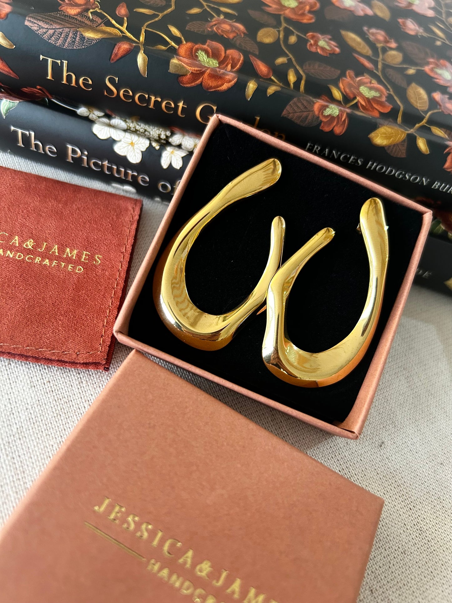 Gold earrings in a box with 'Jessica & James' branding on a textured surface.