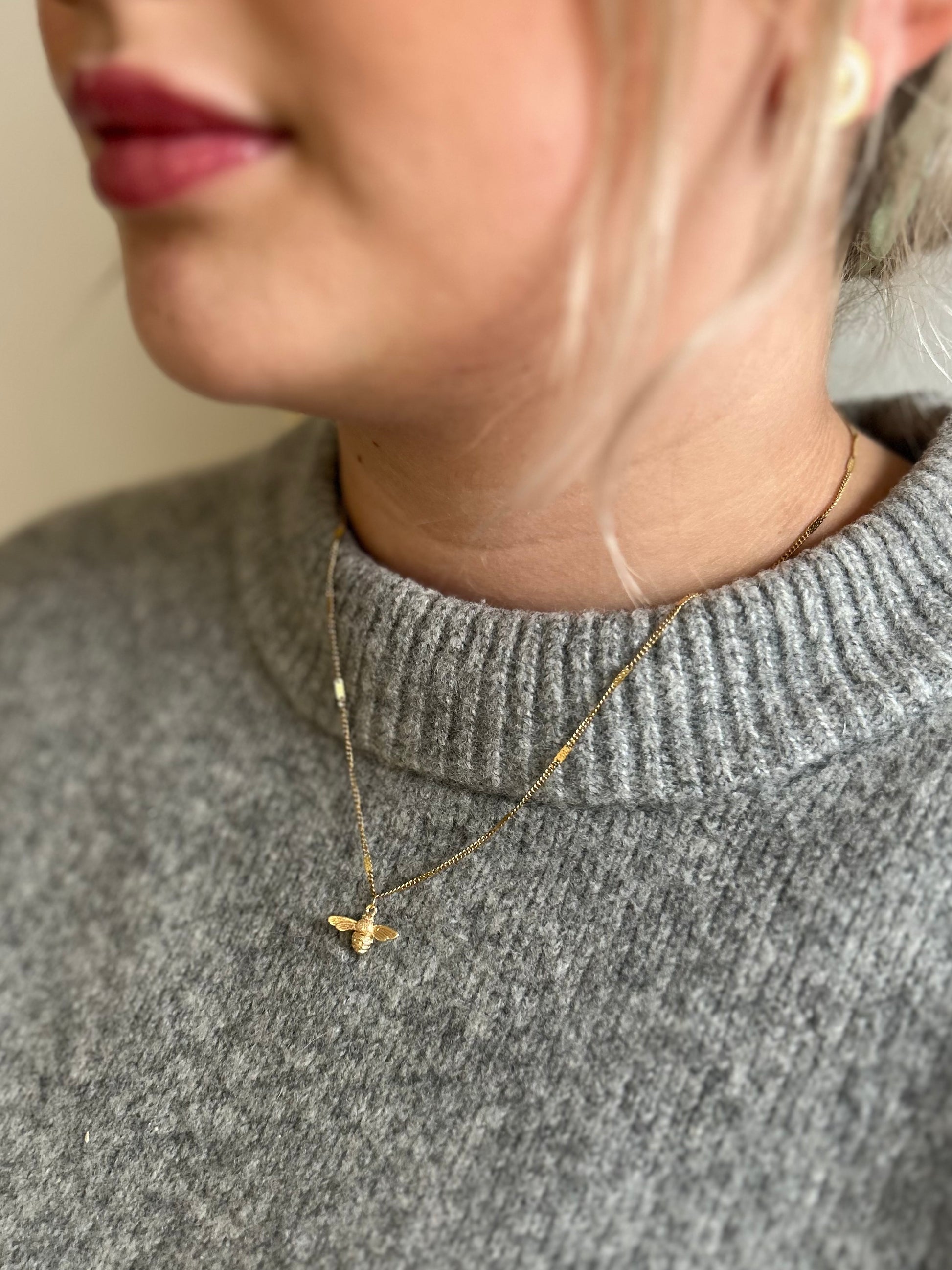 Person wearing a gold bee pendant necklace with a small pendant against a neutral background