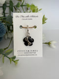 A black floral collar charm with a silver metal connector and keyring, displayed on a white branded card labeled Jessica & James Handcrafted, surrounded by green foliage and white flowers.