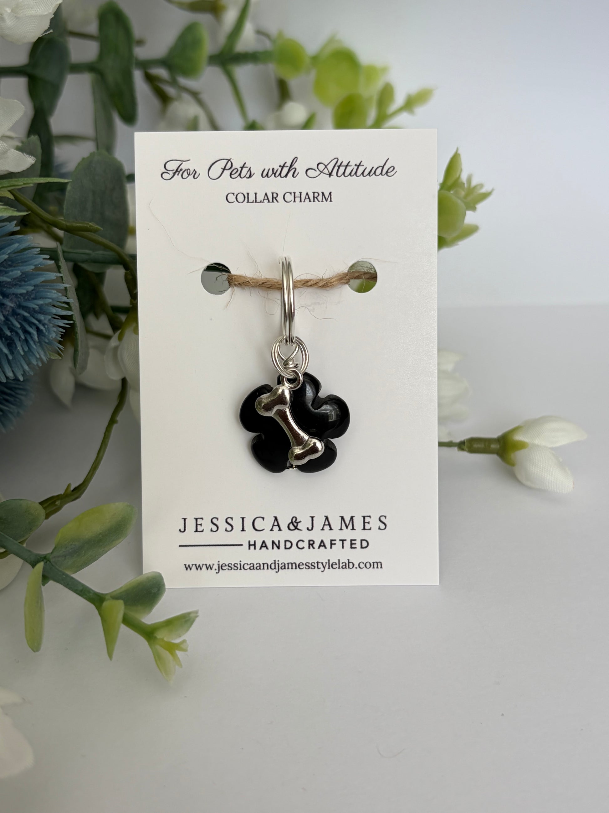 A black floral collar charm with a silver metal connector and keyring, displayed on a white branded card labeled Jessica & James Handcrafted, surrounded by green foliage and white flowers.