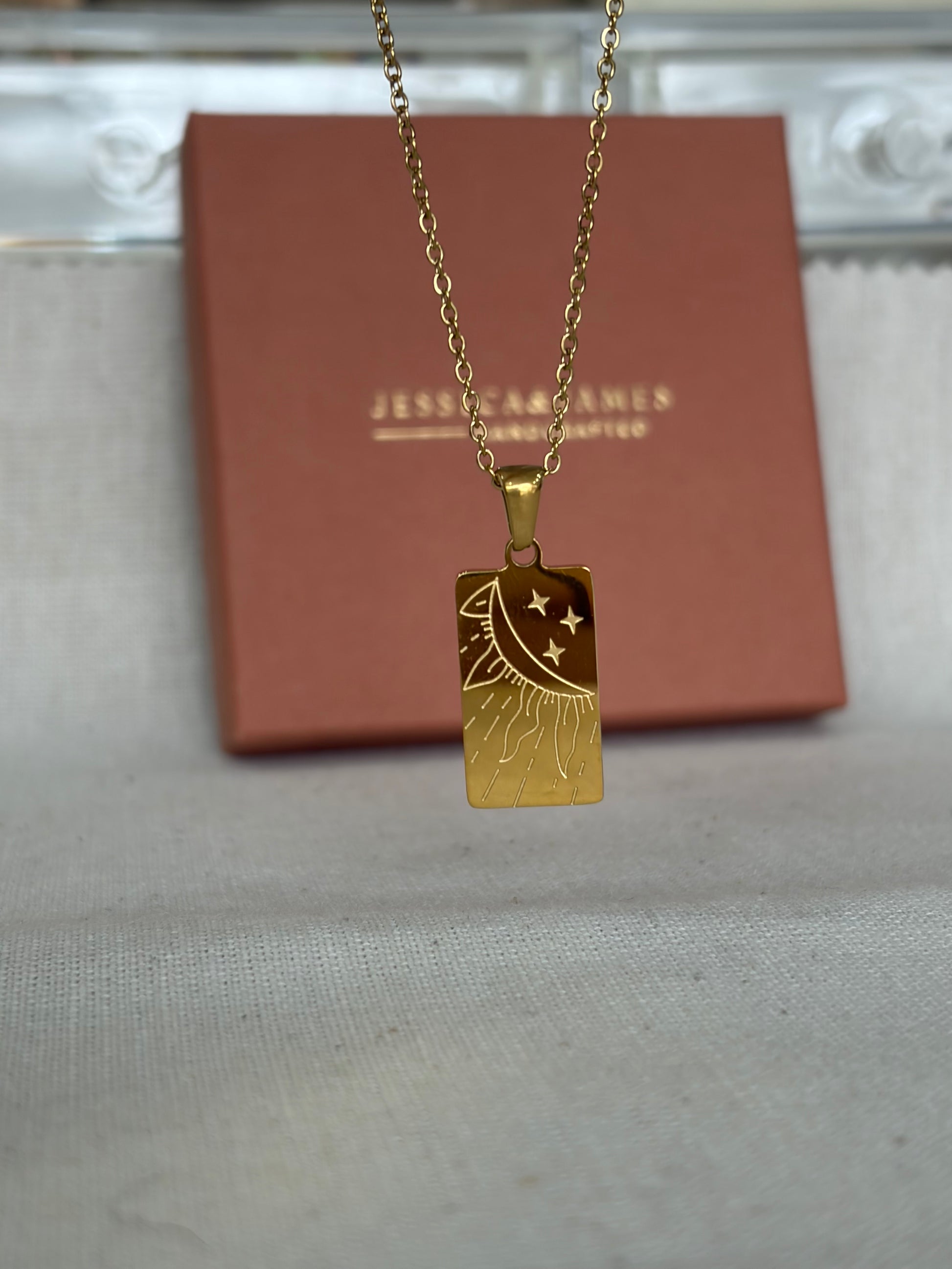 A gold-toned rectangular pendant necklace with an engraved crescent moon and stars design, hanging in front of a brown jewelry box. The box has the brand name 'JESSICA & JAMES ' printed on it.