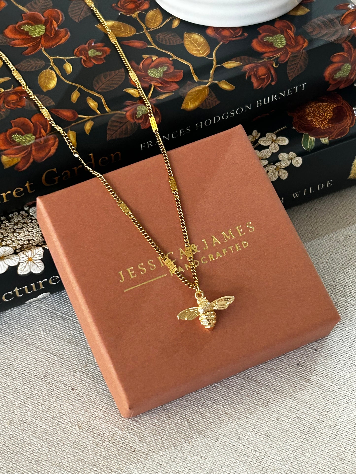 Gold necklace with a bee pendant on a brown box with 'Jessica and James' branding, against a floral book background.