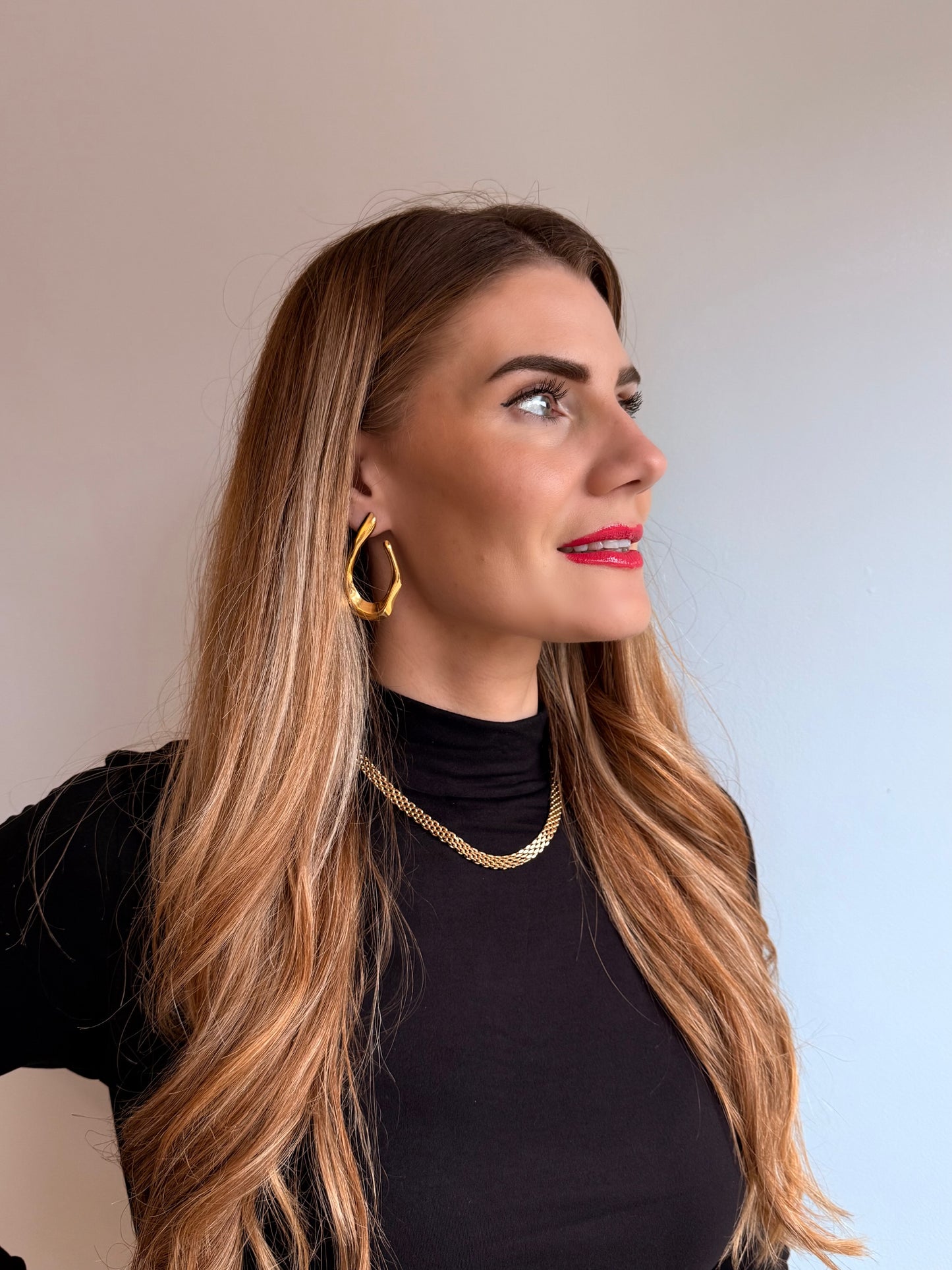 Woman with long blonde hair wearing gold hoop earrings and a black top against a neutral background