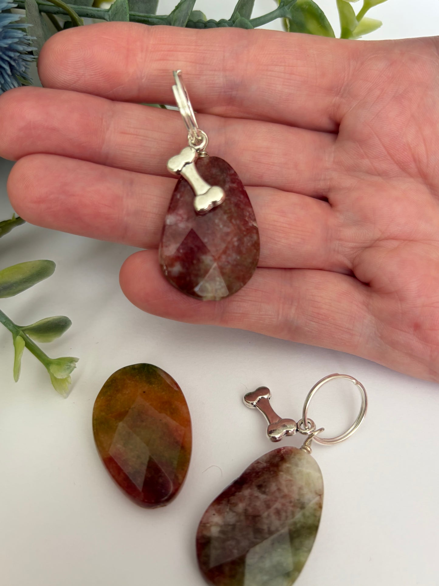 A hand holds a teardrop-shaped faceted gemstone pendant for dogs with a silver bone charm, with two matching gemstone pendants displayed below on a white surface with green leaves in the background.