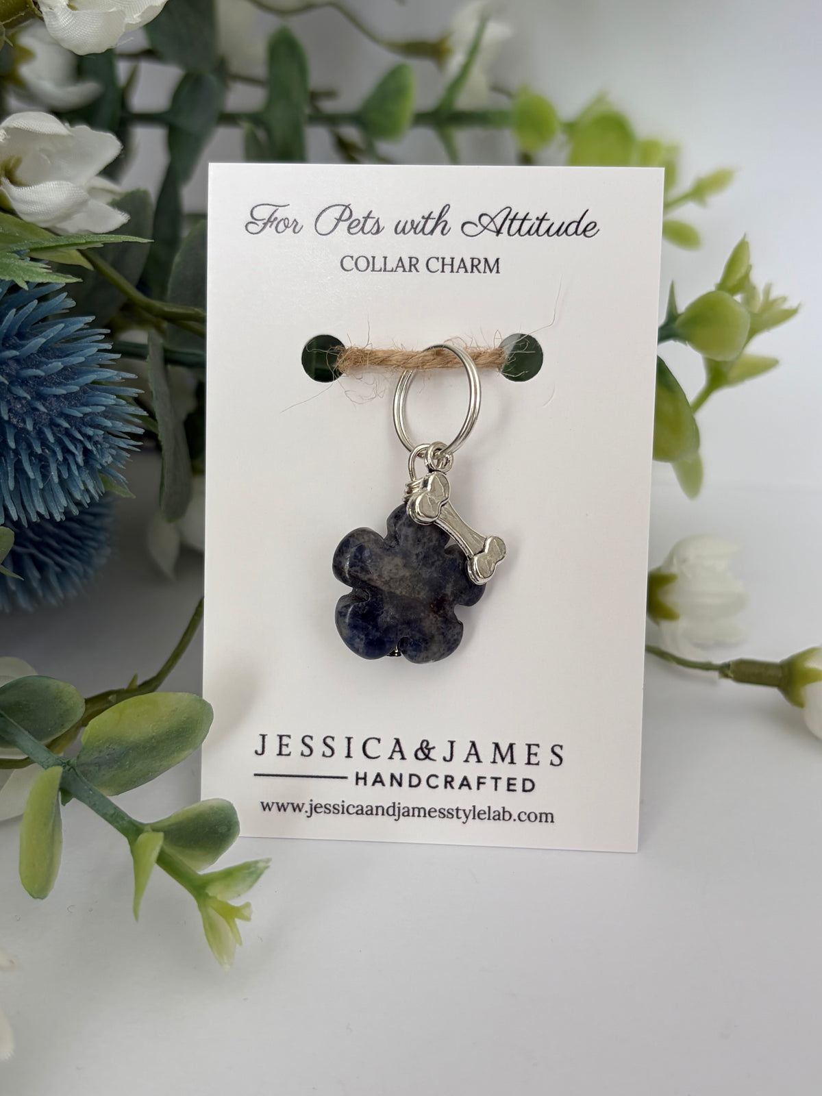 A pet collar charm card featuring a dark blue-gray sodalite flower charm attached to a silver keyring, displayed on a white branded card with decorative greenery in the background.