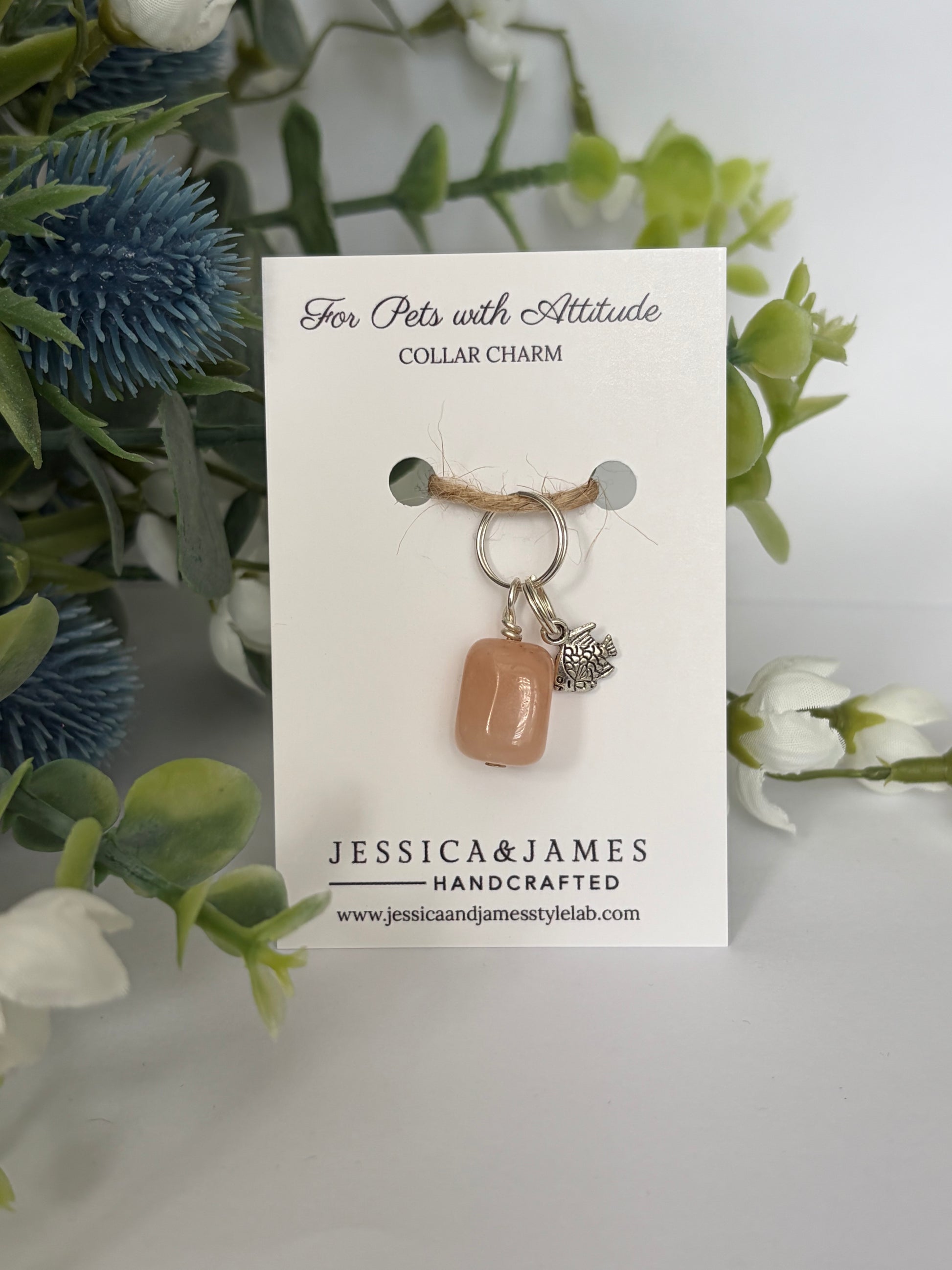 A handmade collar charm for cats by Jessica & James, featuring a peach-colored stone and a silver floral accent, displayed on a white card with decorative green and blue flowers in the background.