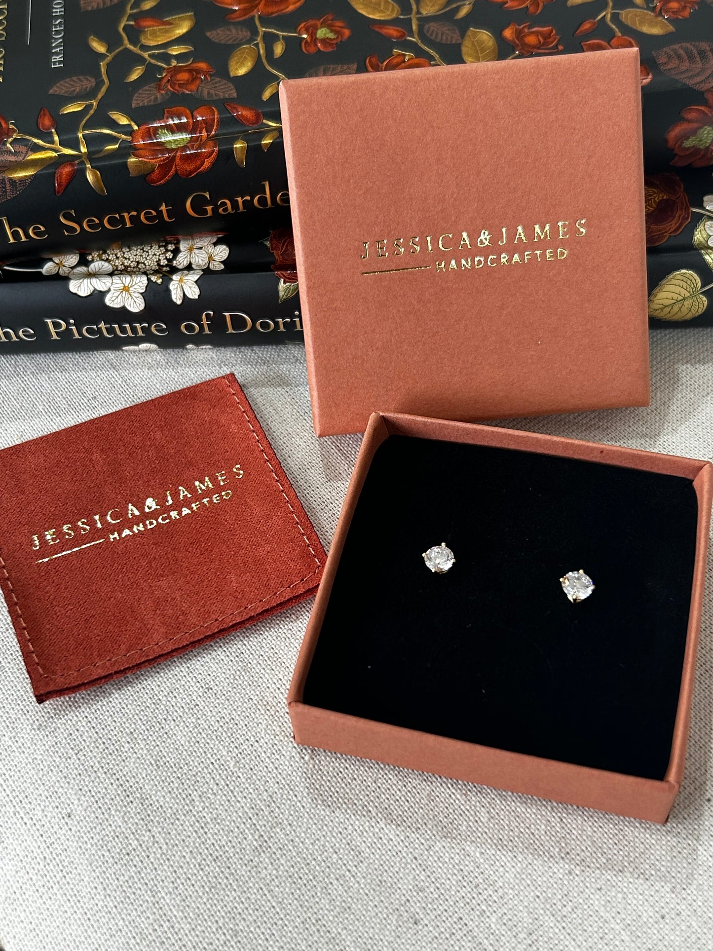 Diamond earrings in a brown jewelry box with 'Jessica James' branding, on a textured surface with books in the background.