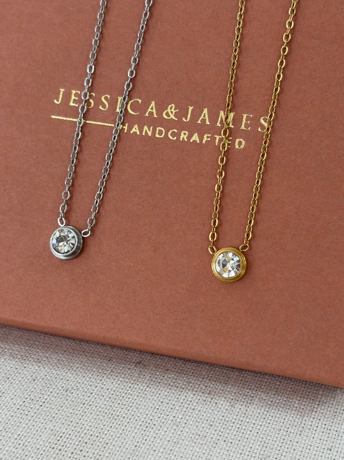 Two delicate necklaces with round bezel-set clear gemstones, one gold-toned and one silver-toned, displayed on a brown presentation box with the brand name 'JESSICA & JAMES HANDCRAFTED'.