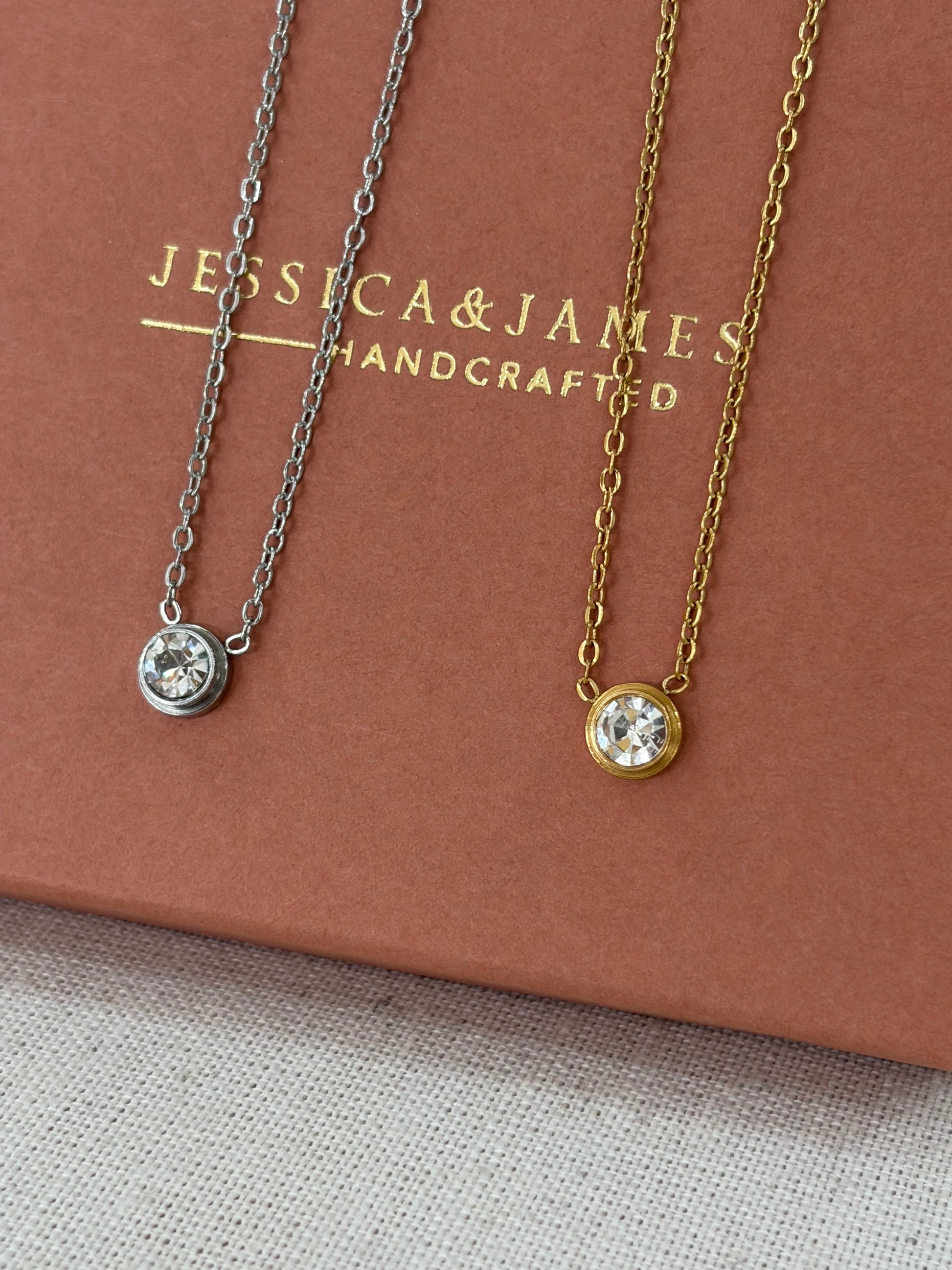 Two delicate necklaces with round bezel-set clear gemstones, one gold-toned and one silver-toned, displayed on a brown presentation box with the brand name 'JESSICA & JAMES HANDCRAFTED'.