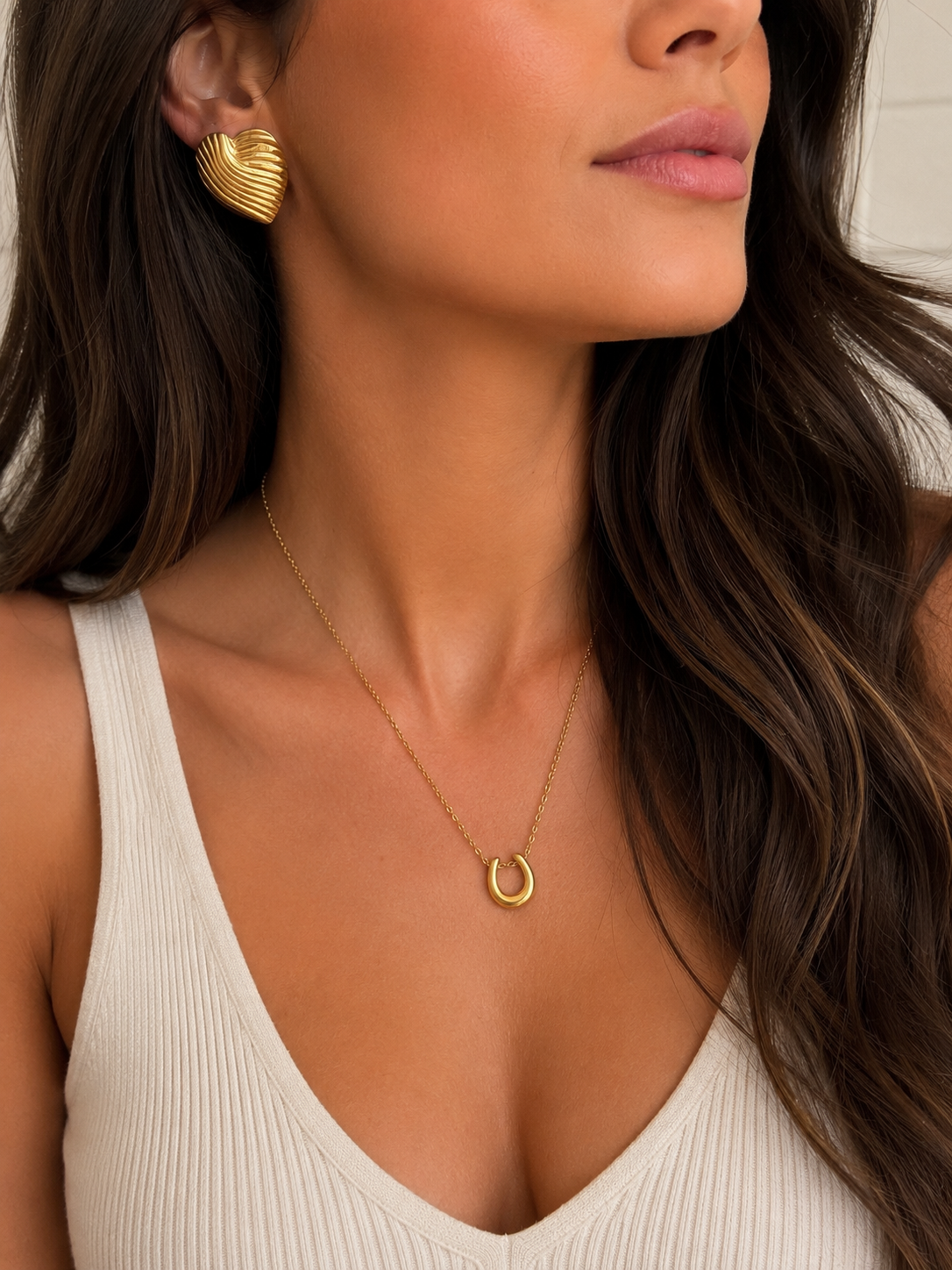A close-up lifestyle shot of a woman wearing a delicate gold crescent moon necklace and a textured gold earring, paired with a cream-colored ribbed tank top. Lucky horse shoe