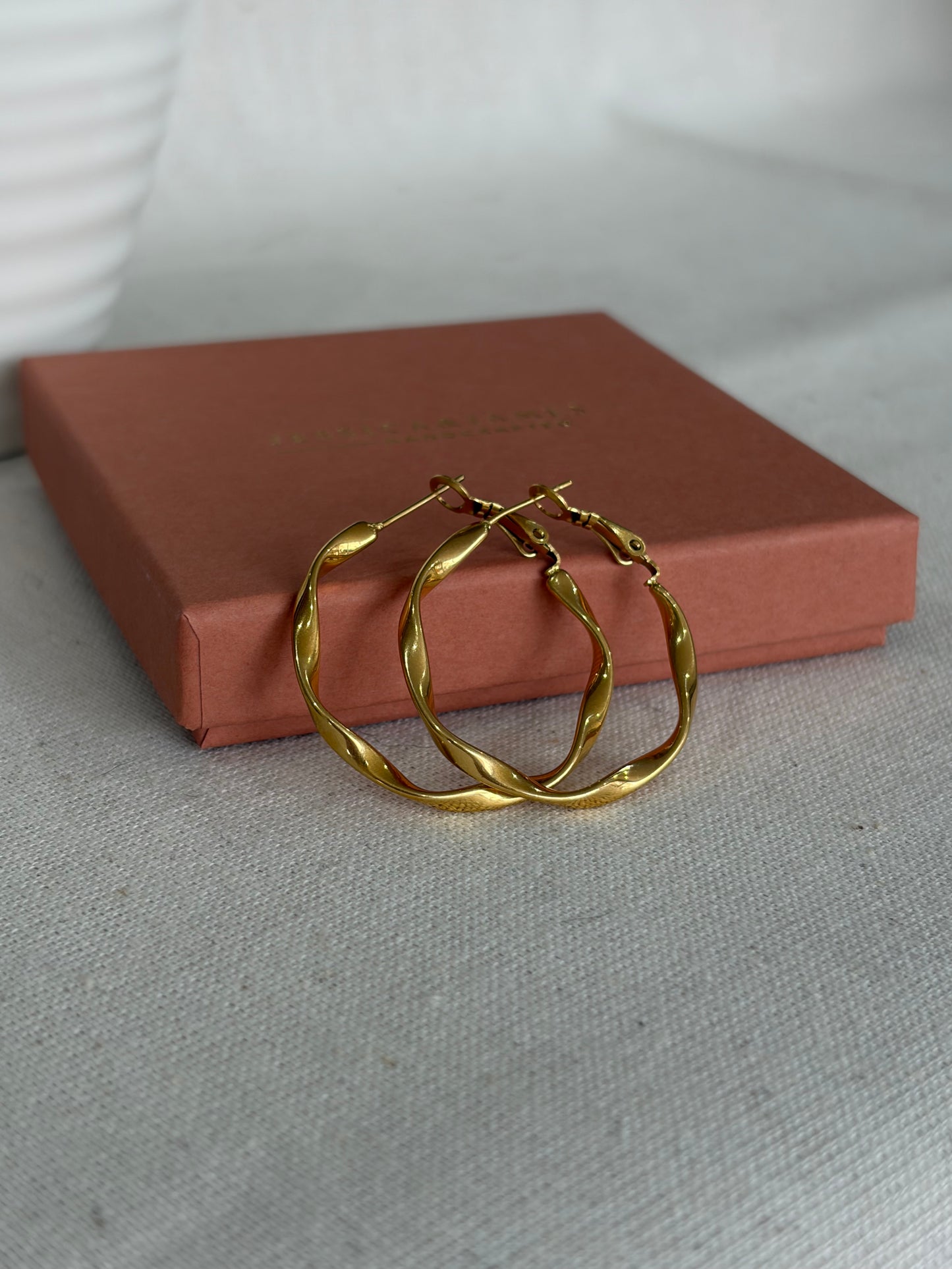 Twisted gold hoop earrings resting on a Jessica and James jewellery box against a light gray textured fabric background.