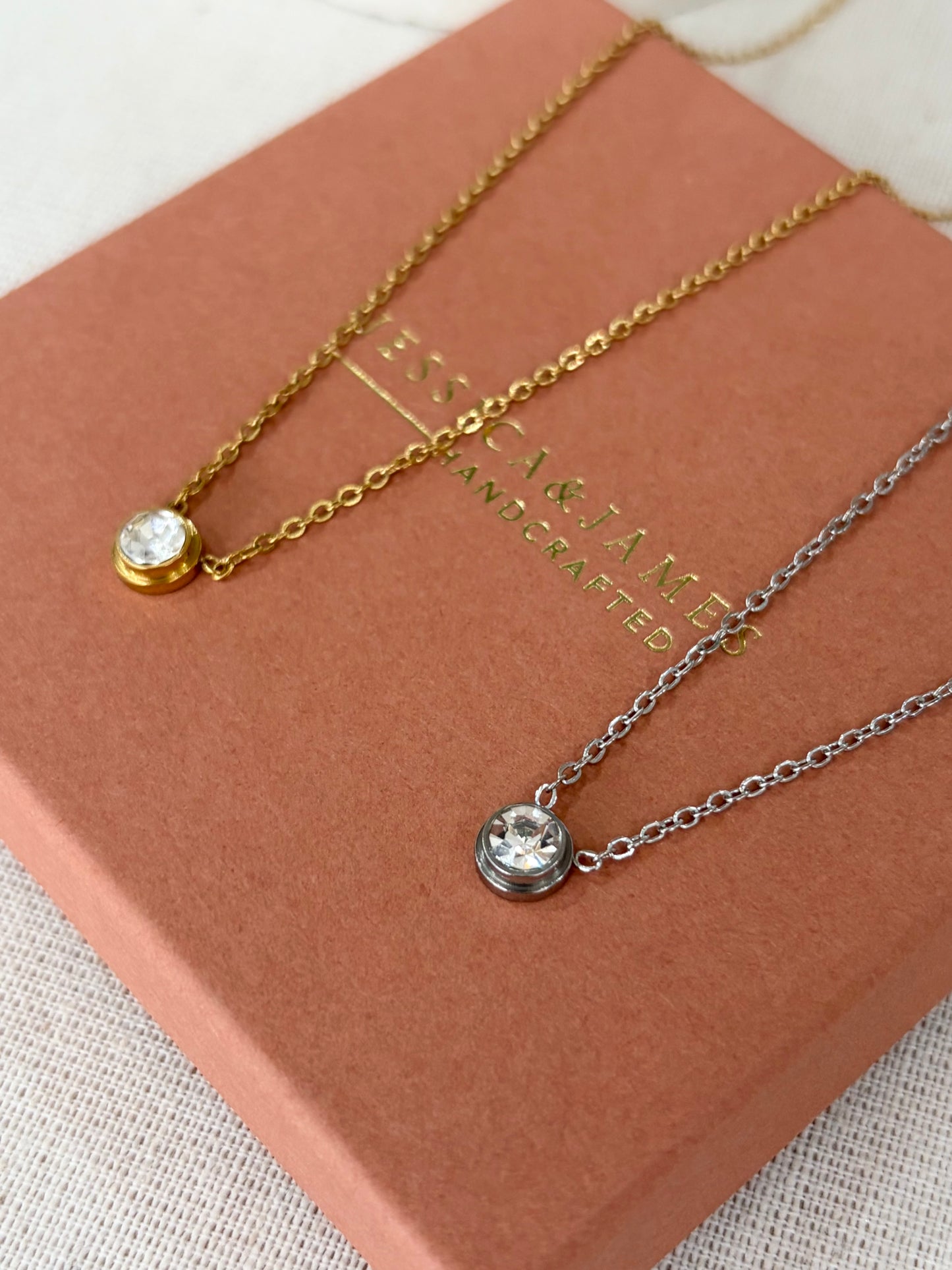 Two delicate round bezel-set crystal necklaces, one gold-toned and one silver-toned, displayed on a dusty rose paper bag with the brand name 'JESSICA & JAMES' printed on it.