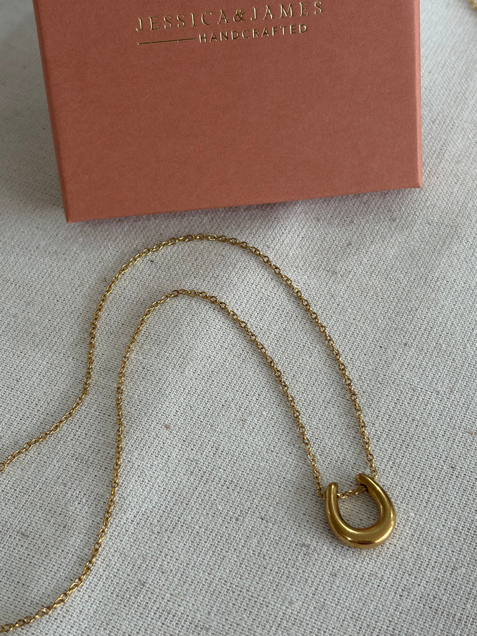 A gold-toned chain necklace with a horseshoe pendant, laid on a light textured fabric next to a dusty pink jewelry box embossed with the brand name Jessica James.