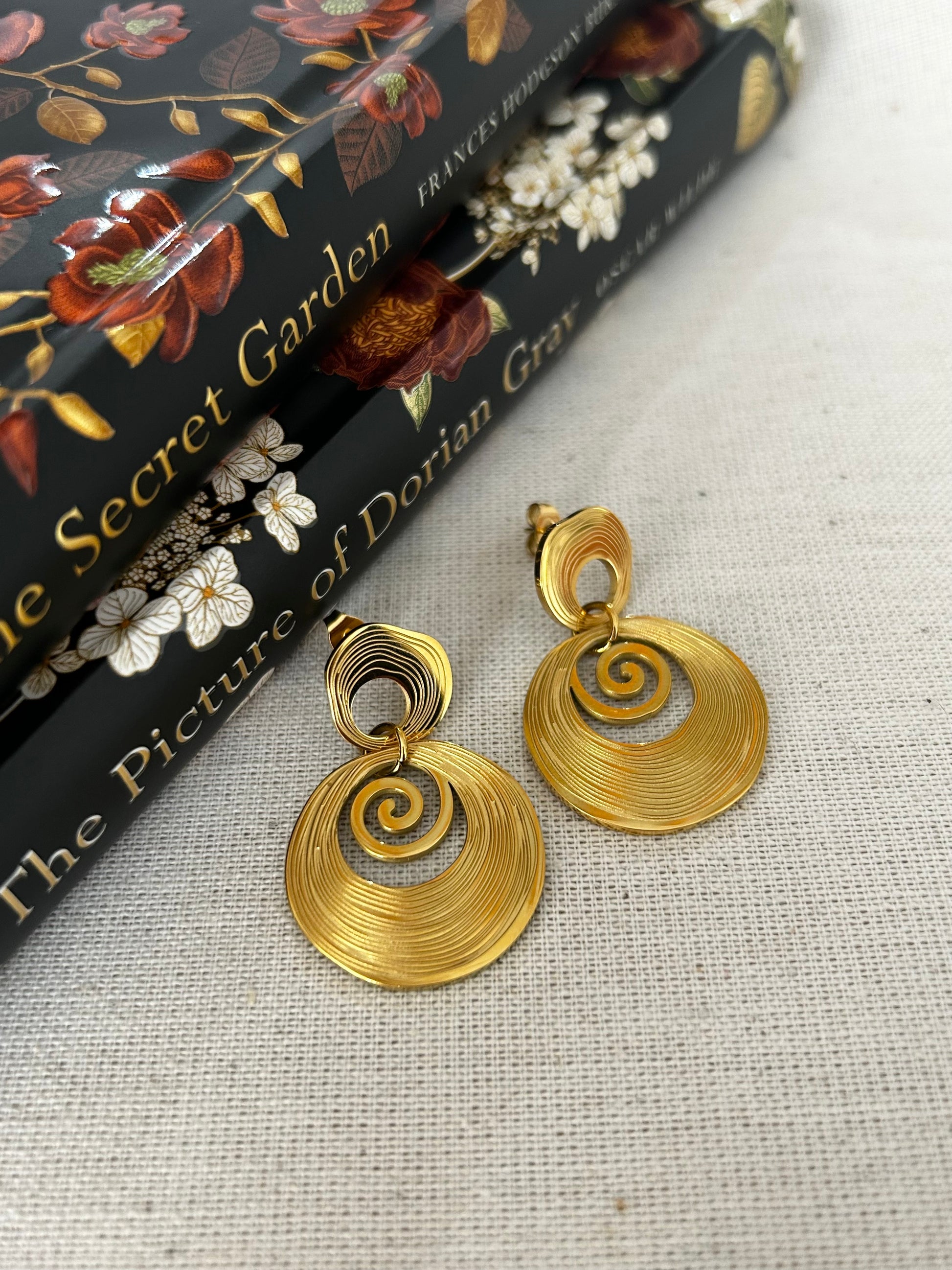 Gold spiral earrings on a textured surface with a book titled 'The Picture of Dorian Gray' in the background.