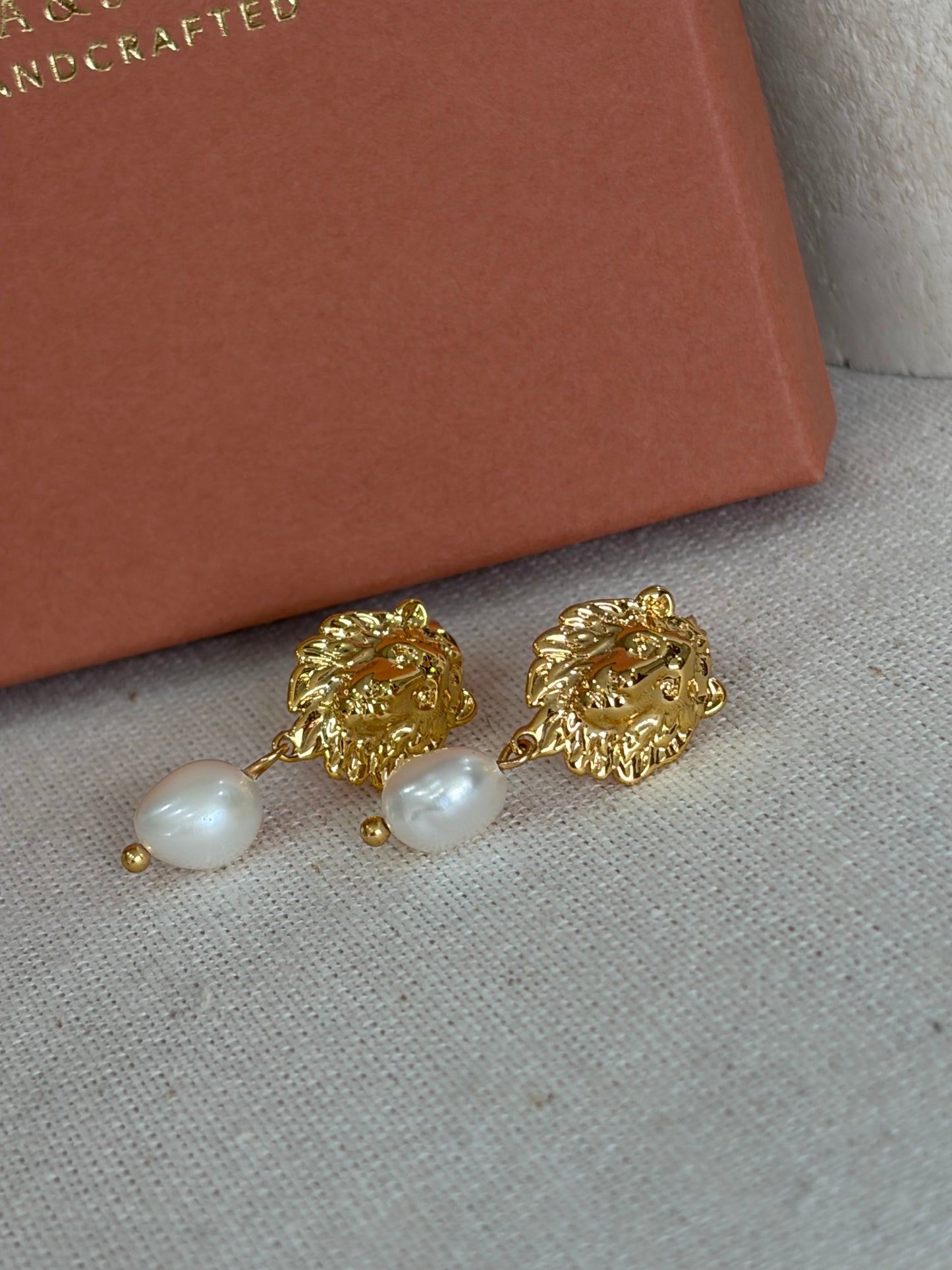 A pair of gold lion-head drop earrings with white pearl drops, displayed on a textured beige fabric surface next to a brown box.