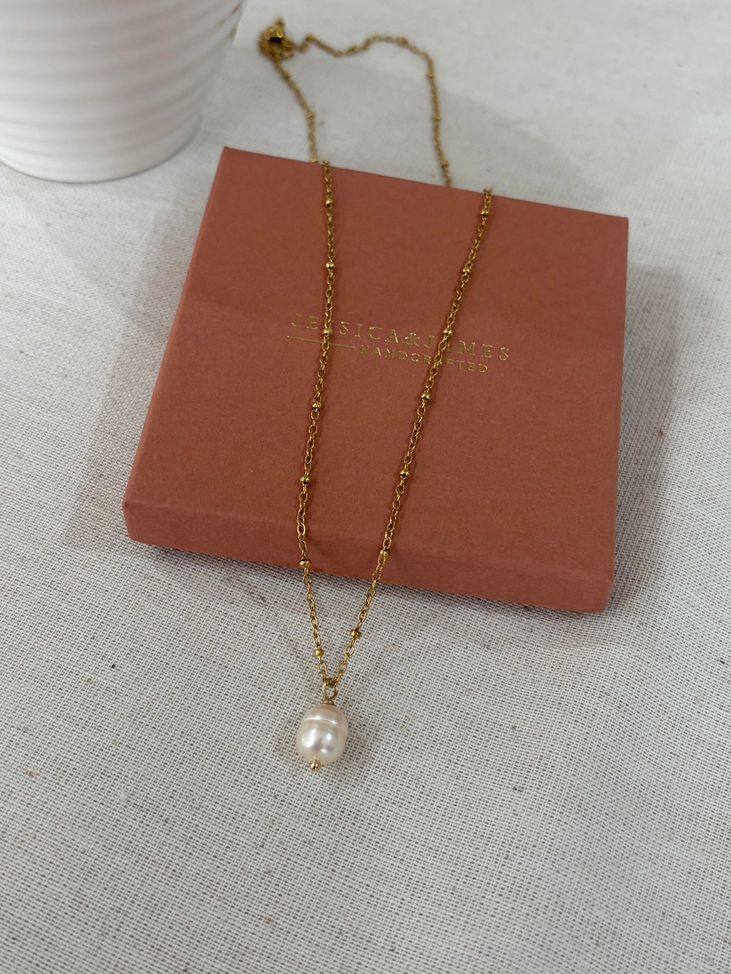 A gold chain necklace with a single white baroque pearl pendant, draped over a terracotta-colored jewelry box on a textured light gray fabric surface.