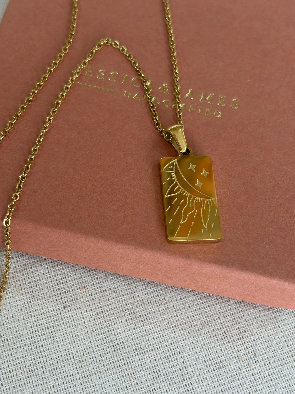 A gold-tone rectangular pendant necklace with an engraved sun and moon design, resting on a dusty pink jewelry box. The box has the brand name 'JESSICA & JAMES  JEWELLERY ' embossed on it.