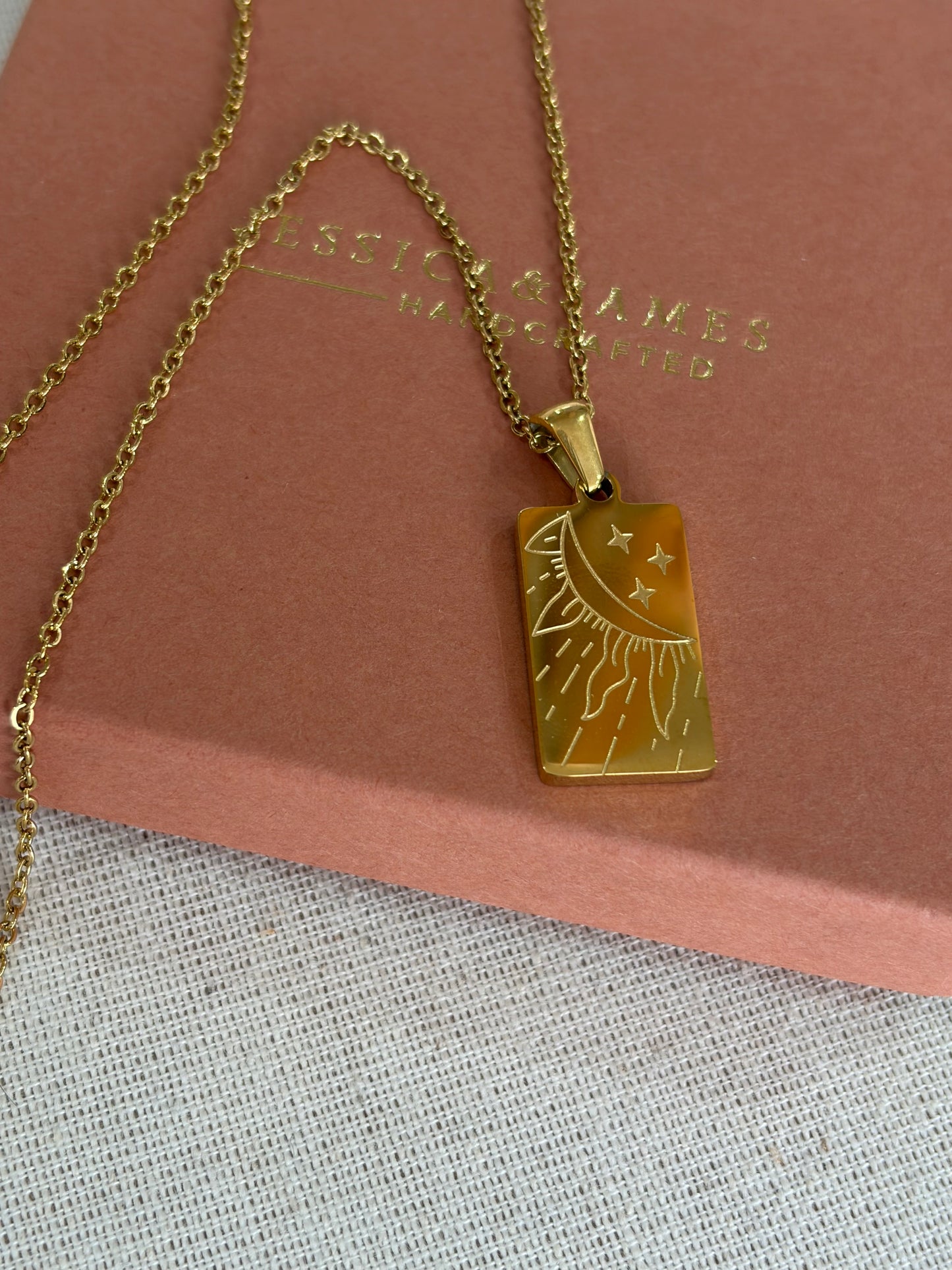 A gold-tone rectangular pendant necklace with an engraved sun and moon design, resting on a dusty pink jewelry box. The box has the brand name 'JESSICA & JAMES  JEWELLERY ' embossed on it.