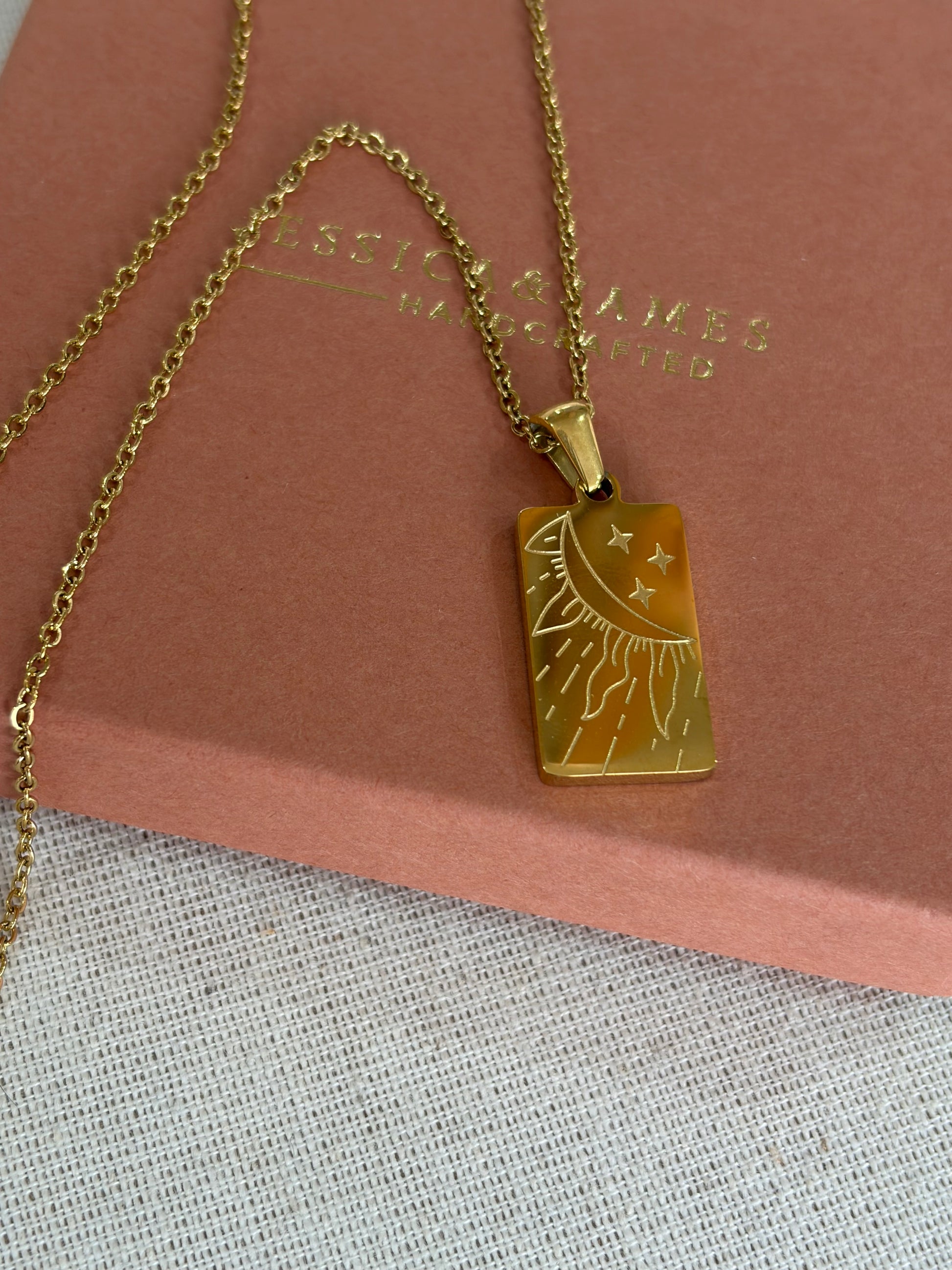 A gold-tone rectangular pendant necklace with an engraved sun and moon design, resting on a dusty pink jewelry box. The box has the brand name 'JESSICA & JAMES  JEWELLERY ' embossed on it.
