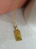 A gold rectangular pendant necklace with a sun and star design, hanging from a gold chain on a textured beige fabric surface.