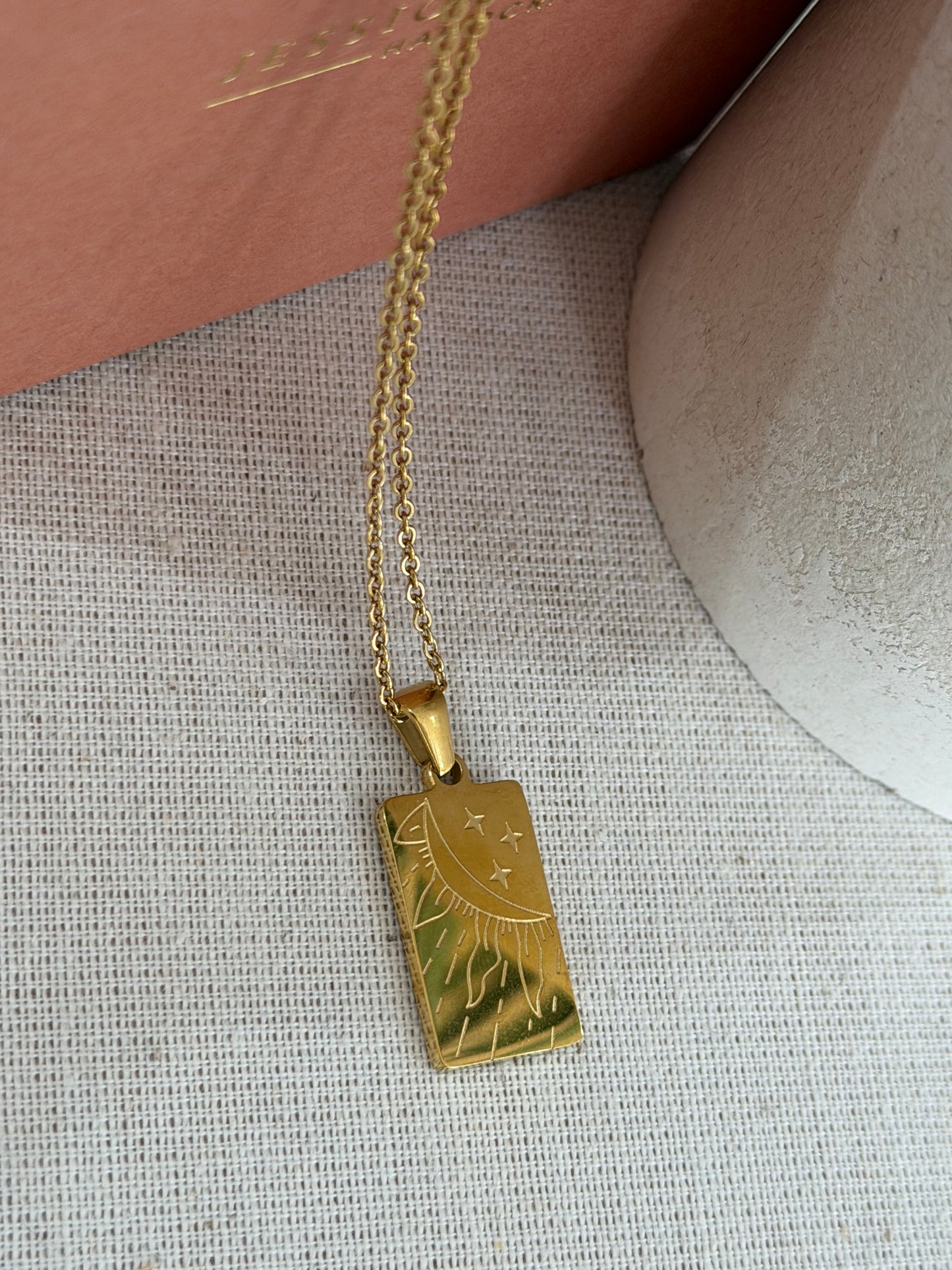 A gold rectangular pendant necklace with a sun and star design, hanging from a gold chain on a textured beige fabric surface.