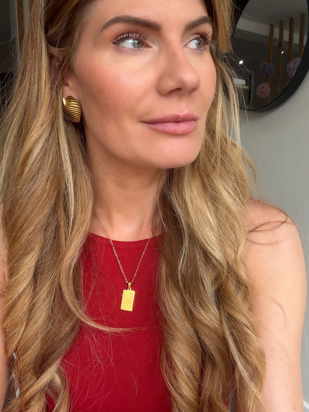 A close-up lifestyle shot of a woman with long wavy blonde hair wearing a gold rectangular pendant necklace and a red top.