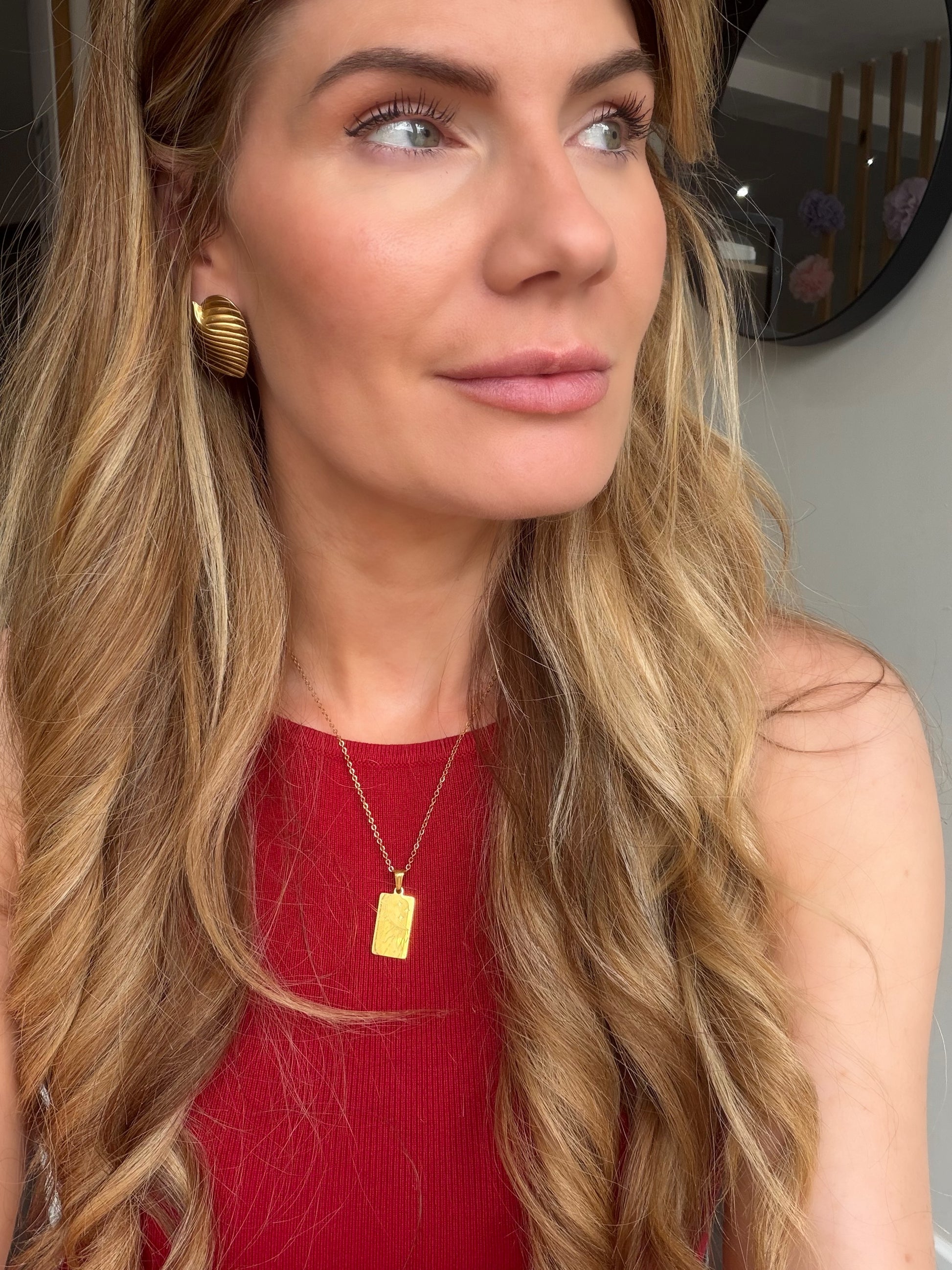 A close-up lifestyle shot of a woman with long wavy blonde hair wearing a gold rectangular pendant necklace and a red top.