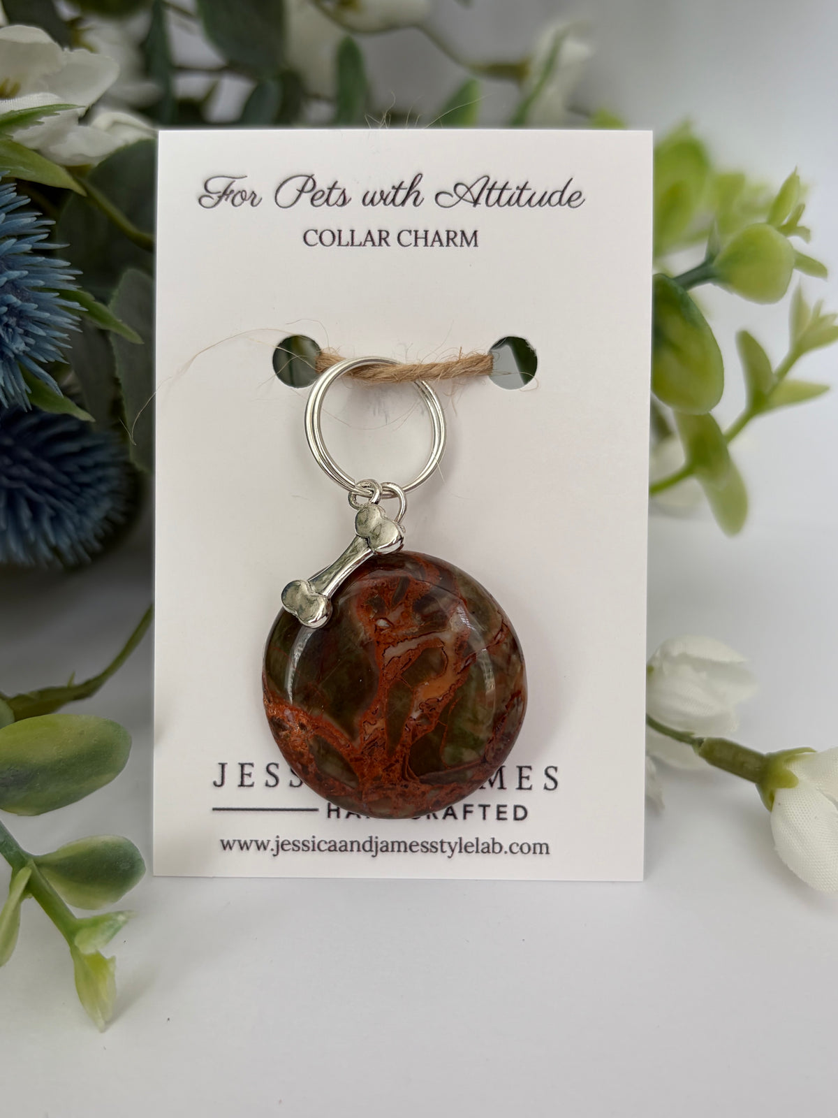 A pet collar charm displayed on a white branded Jessica and James pets card, featuring an oval marbled jasper orange pendant with a small silver bone accent and silver ring, surrounded by soft green and white floral props.