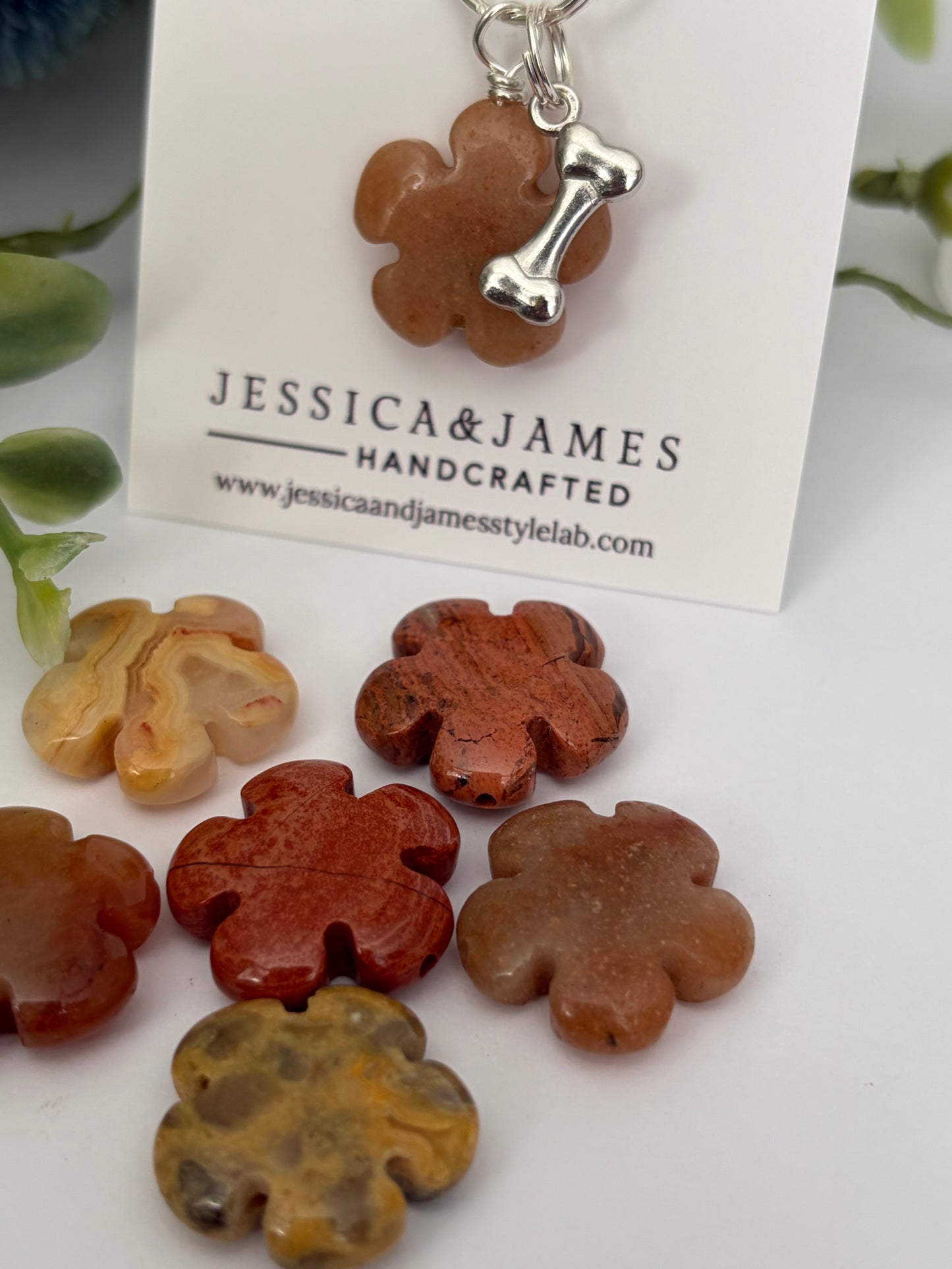 A set of handcrafted pet memorial jewelry pendants shaped like paw prints and bone charms in earthy stone colors, displayed on a white card labeled 'Jessica & James Handcrafted'.