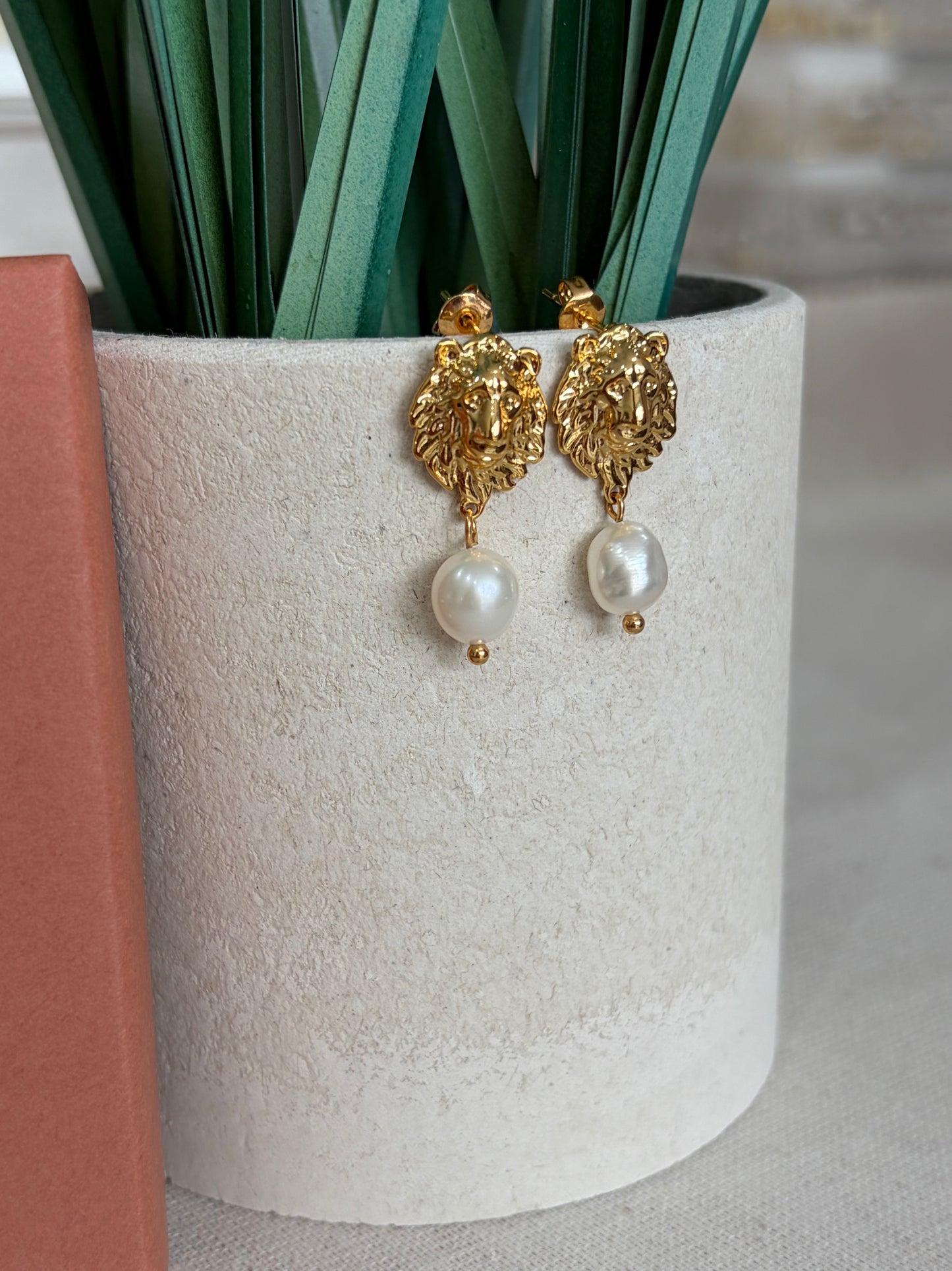 A pair of gold-toned drop earrings with ornate leaf-shaped charms and white pearl beads, displayed on a textured cream cylindrical stand.