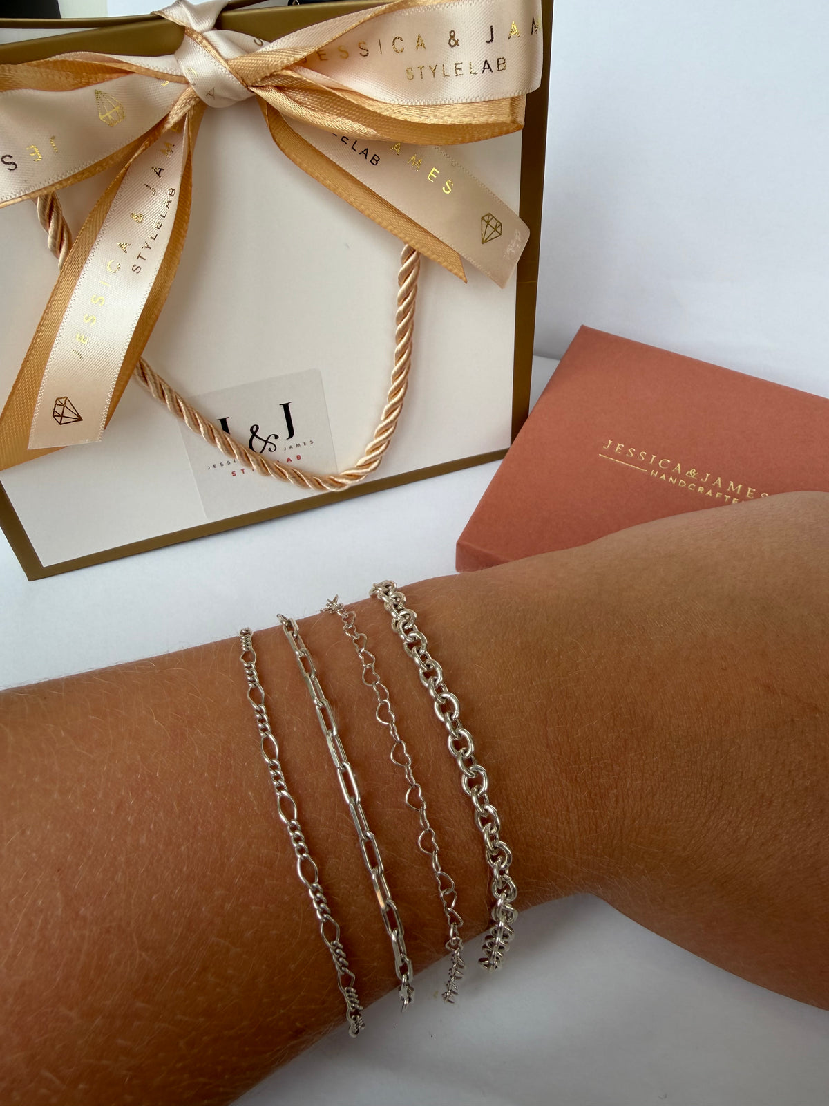A close-up of a person's wrist wearing four sterling silver clasp free permanent bracelets, shown next to a gift box with a cream ribbon printed with the brand name 'Jessica & James Stylelab'.