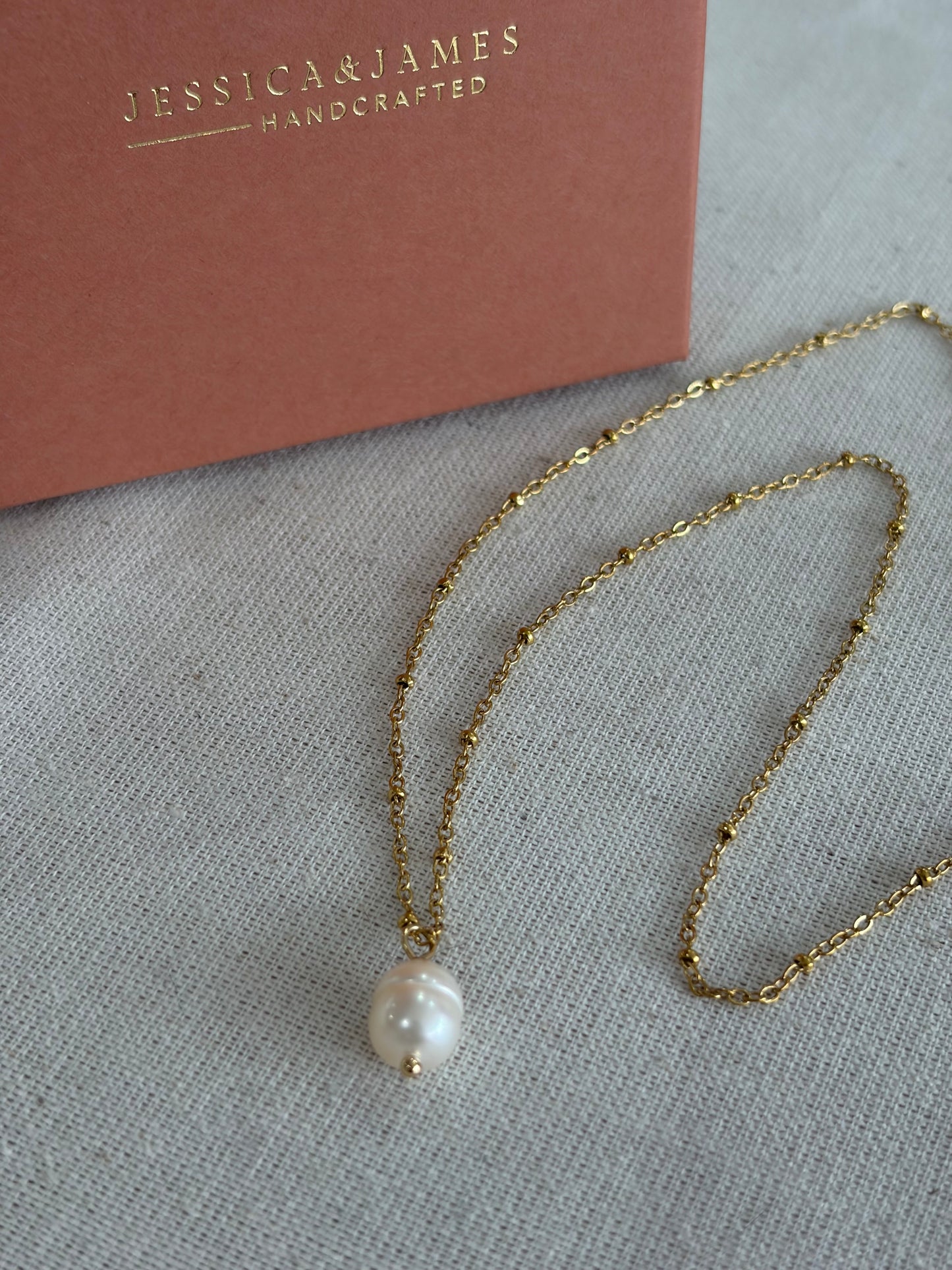 A delicate gold chain necklace with a single white pearl pendant, displayed on a textured light gray fabric next to a dusty rose card printed with the brand name 'JESSICA & JAMES HANDCRAFTED'.