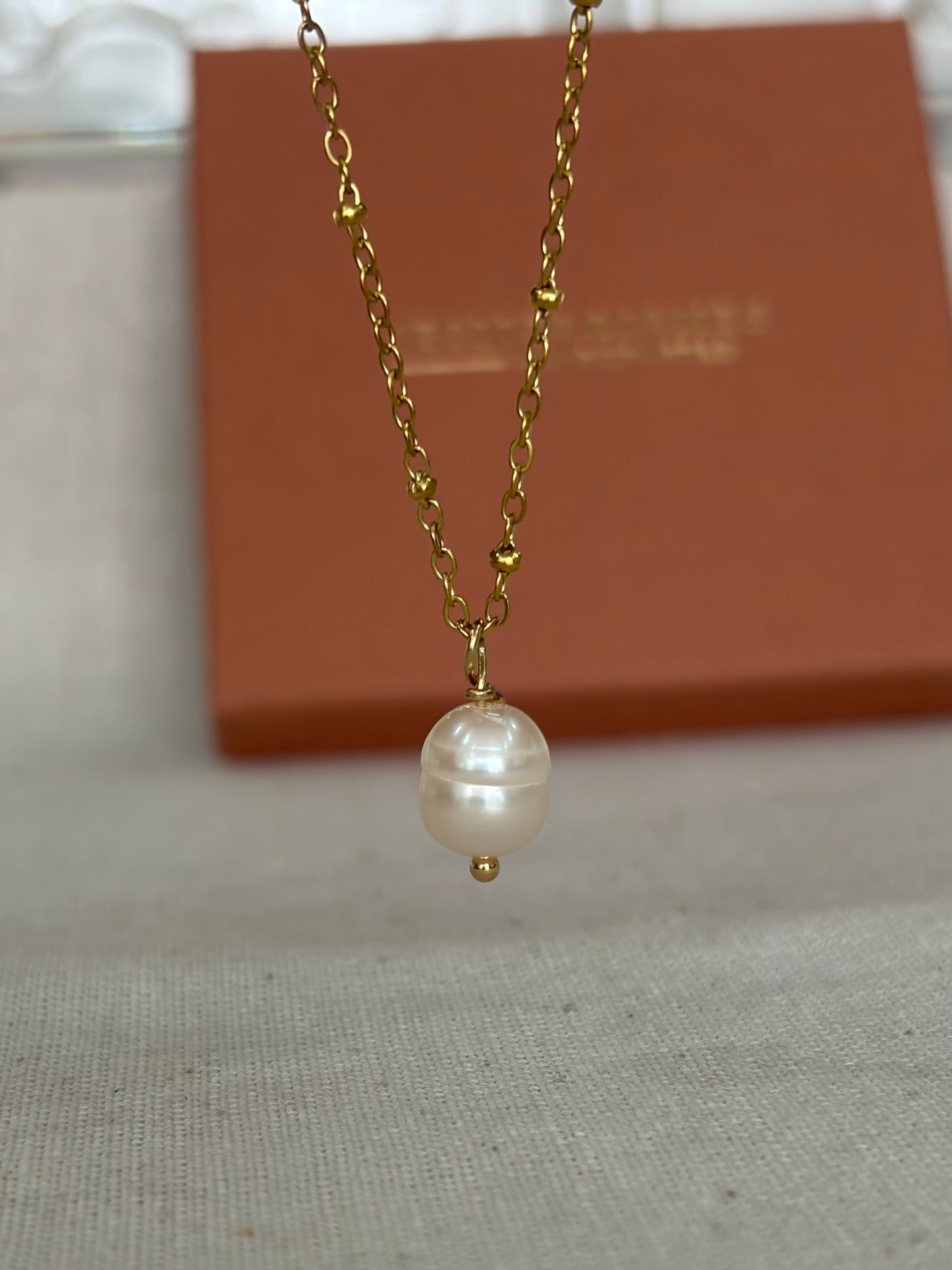 A gold chain necklace featuring a single, irregular white pearl pendant, displayed against a neutral beige fabric background with a brown presentation box in the background.