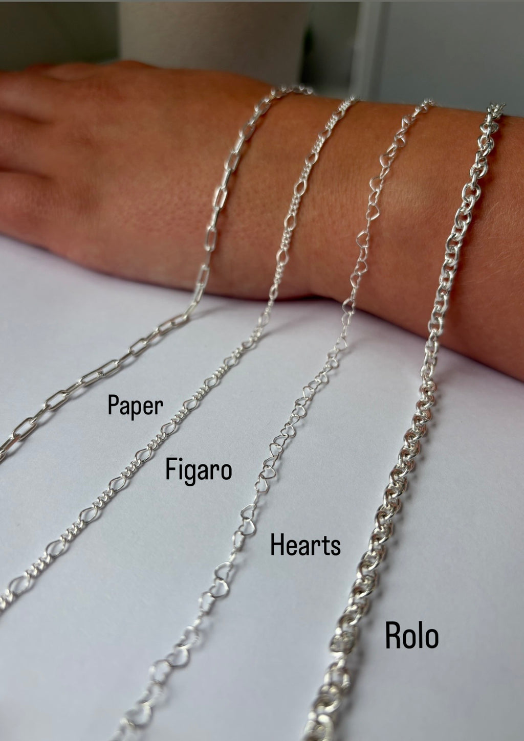 Four sterling silver permanent necklaces labeled 'Paper', 'Figaro', 'Hearts', and 'Rolo' draped over a person's wrist and hand against a light background.