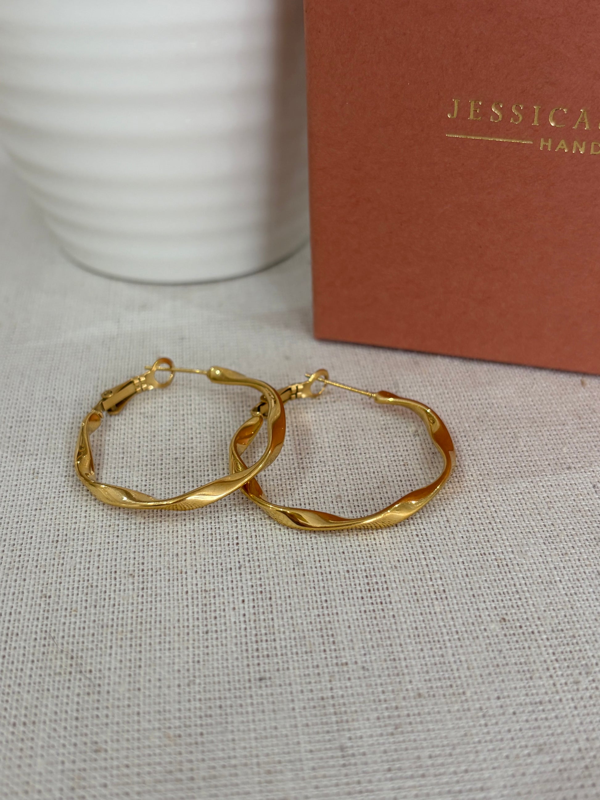 Twisted gold hoop earrings displayed on a textured beige fabric surface, with a white ribbed ceramic cup and a brown paper box in the background. The box features the brand name 'JESSICA  AND JAMES STYLELAB' in gold foil.