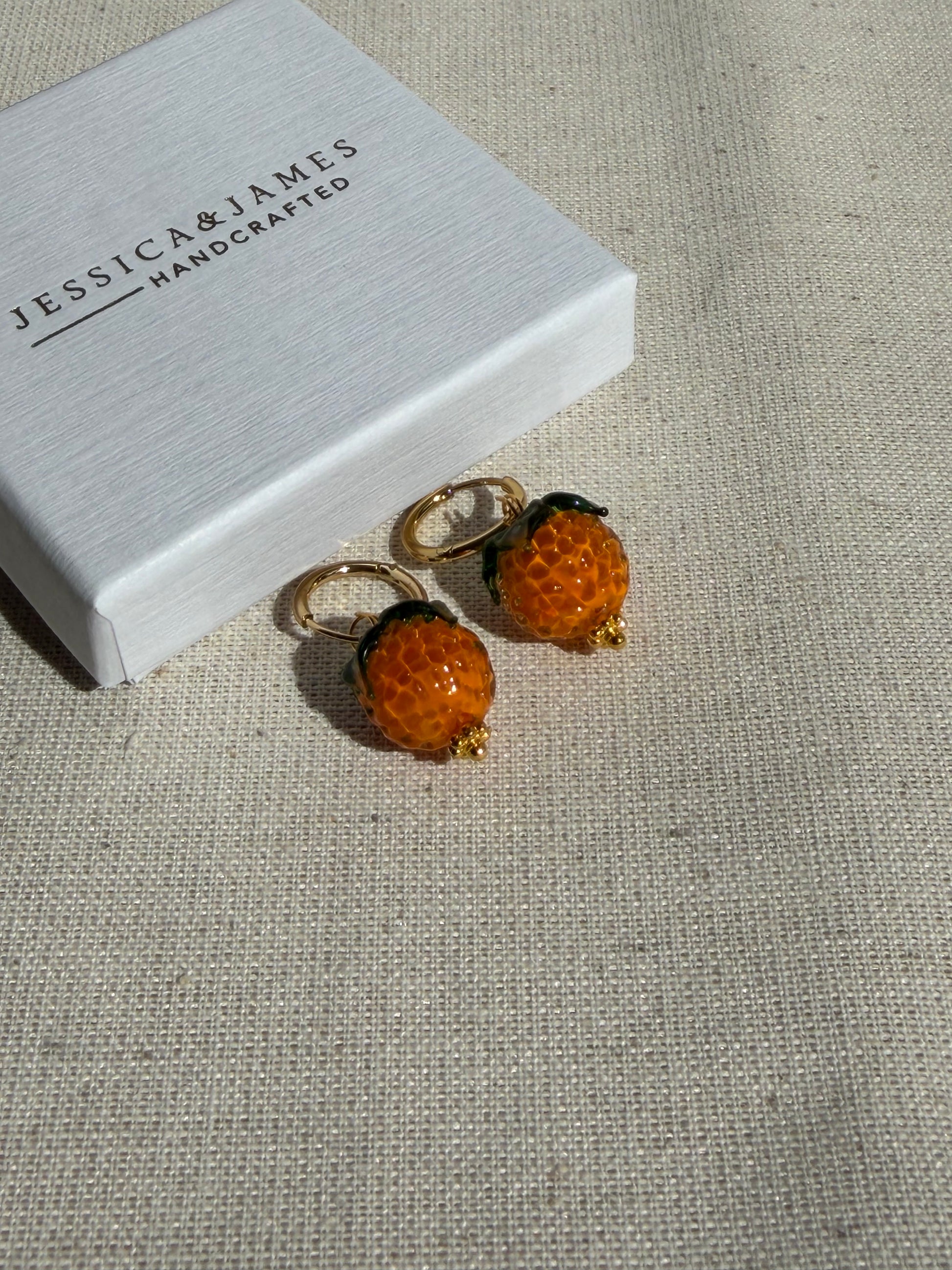 Pair of orange floral earrings on a beige fabric background with 'Jessica James' branding.