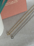 Two delicate chain necklaces, one gold and one silver, each featuring a single bezel-set clear gemstone, displayed on a textured gray fabric next to a dusty pink jewelry box.
