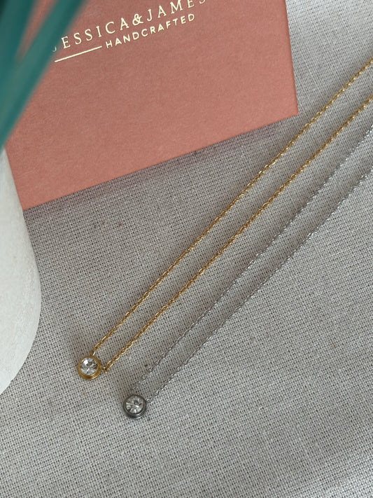 Two delicate chain necklaces, one gold and one silver, each featuring a single bezel-set clear gemstone, displayed on a textured gray fabric next to a dusty pink jewelry box.