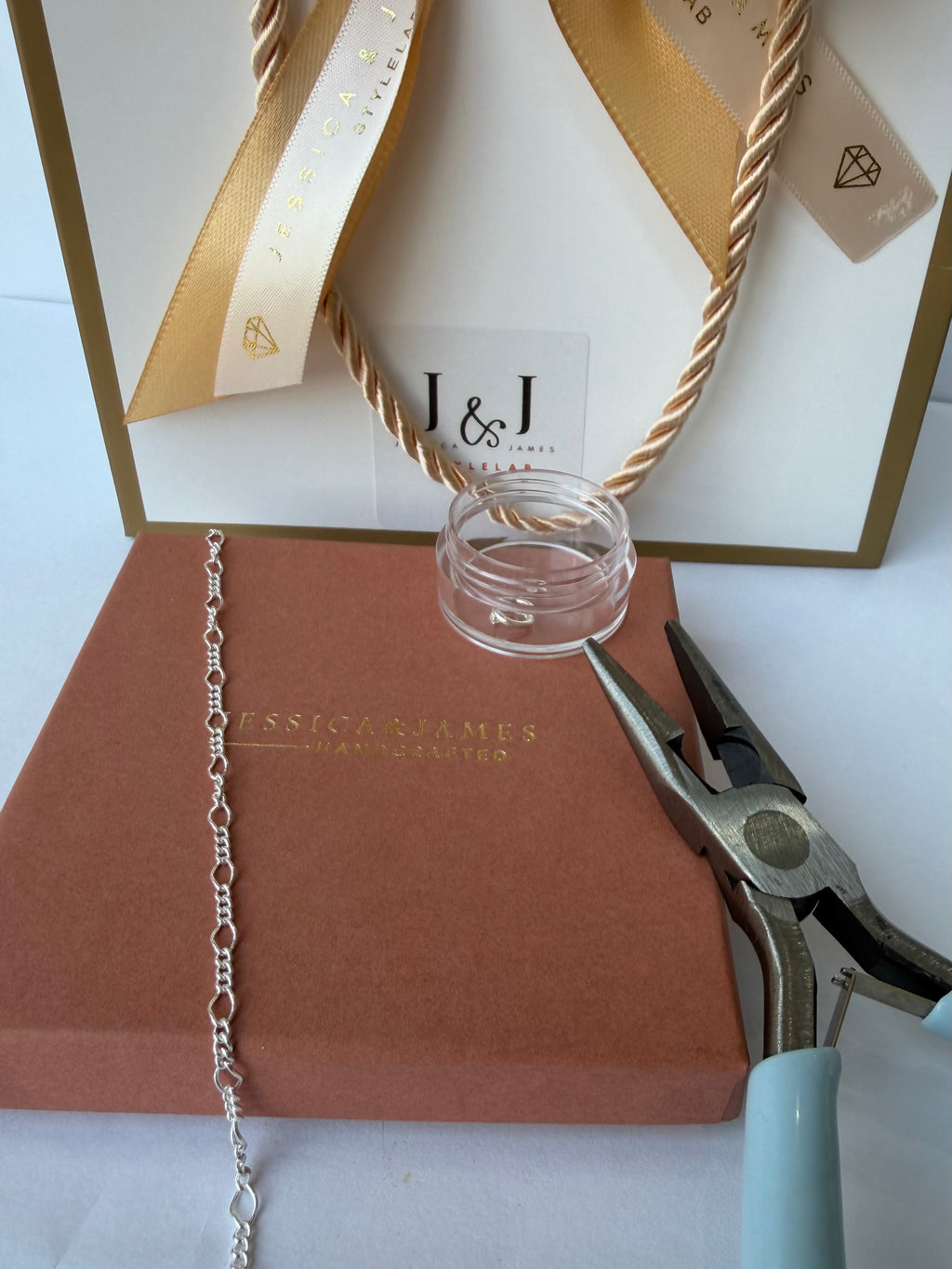 A close-up of permanent jewellery making supplies and packaging, including a silver chain draped over a brown box, a small clear glass jar, metal pliers, and a white gift bag with gold ribbon and the J&J logo.
