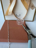 A close-up of permanent jewellery making supplies and packaging, including a silver chain draped over a brown box, a small clear glass jar, metal pliers, and a white gift bag with gold ribbon and the J&J logo.