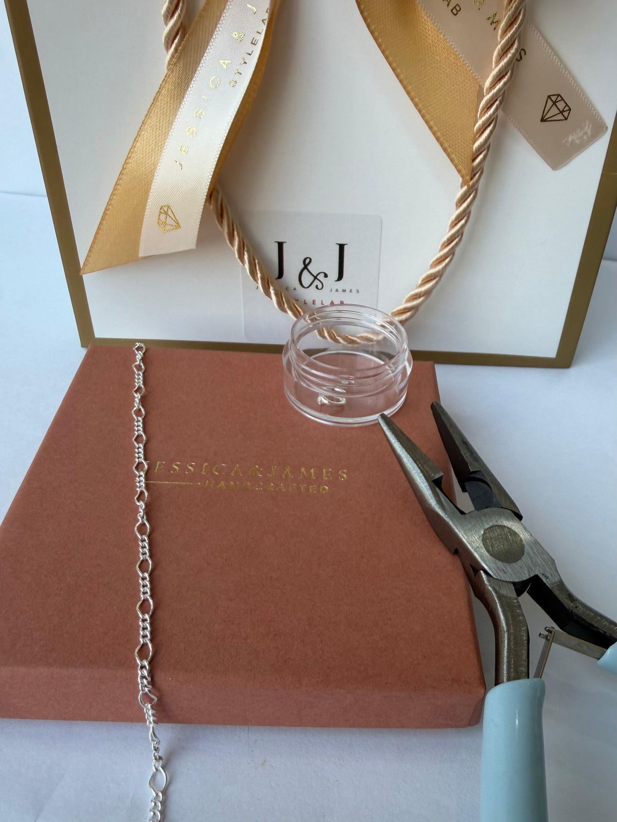 A close-up of permanent jewellery making supplies and packaging, including a silver chain draped over a brown box, a small clear glass jar, metal pliers, and a white gift bag with gold ribbon and the J&J logo.