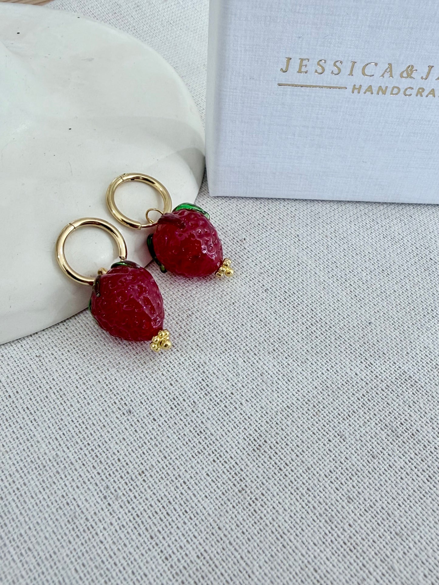 Strawberry-shaped earrings on a textured surface with 'Jessica & James' branding.