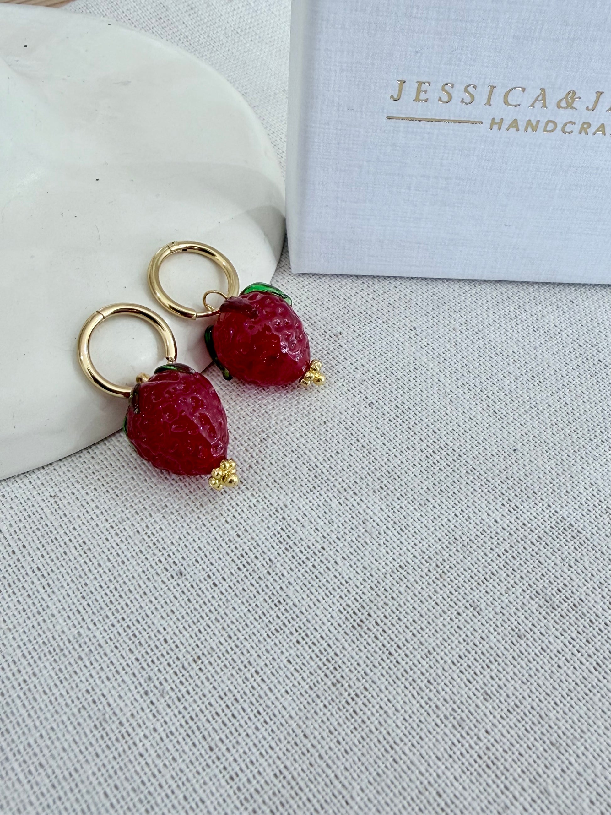 Strawberry-shaped earrings on a textured surface with 'Jessica & James' branding.