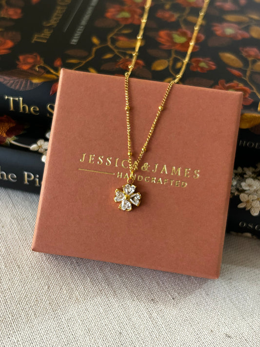 Gold necklace with 'Jessica James' branding, placed on a textured surface.