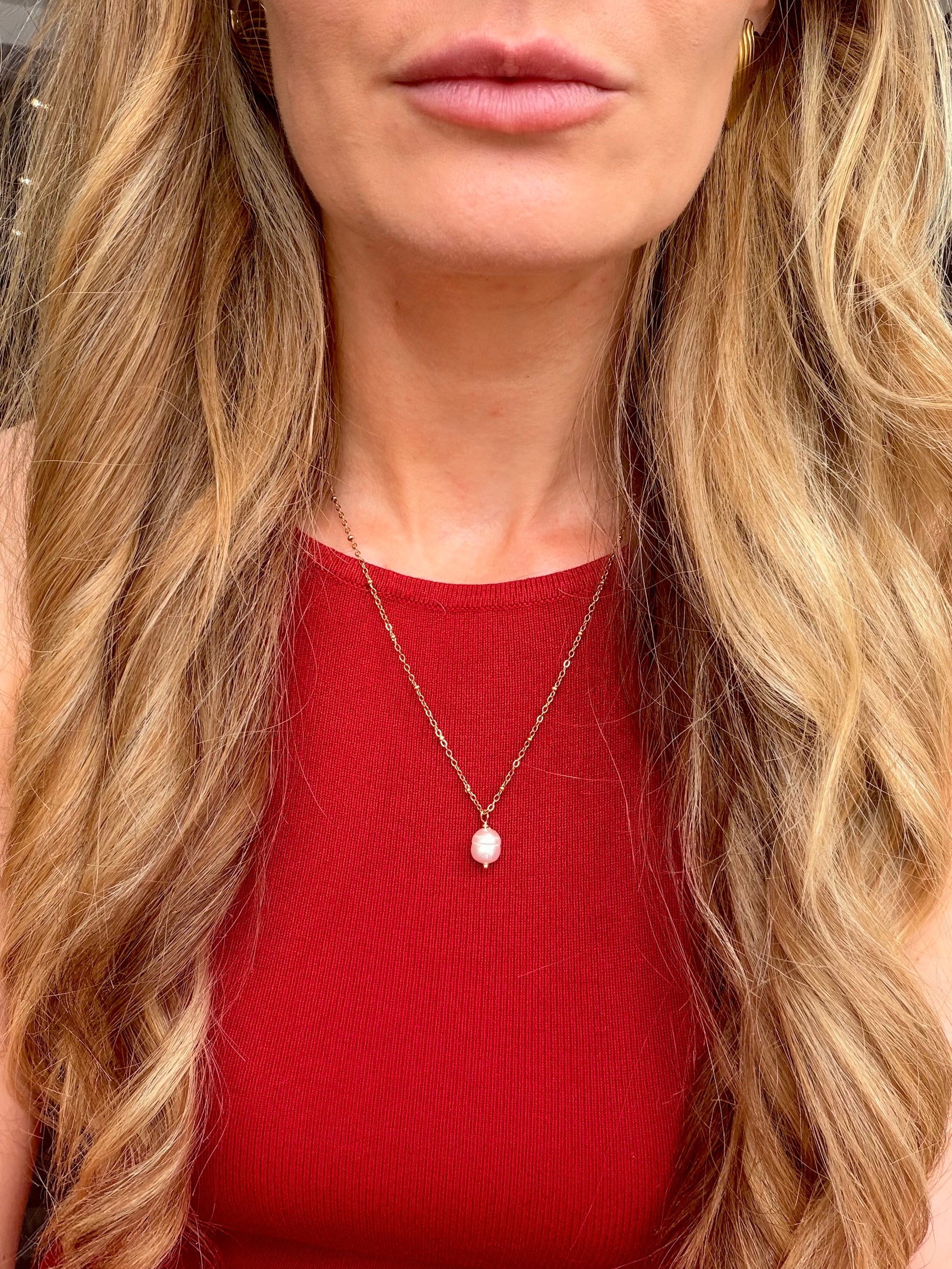 A close-up of a woman wearing a delicate gold chain necklace with a small white pearl pendant, paired with a red ribbed top.