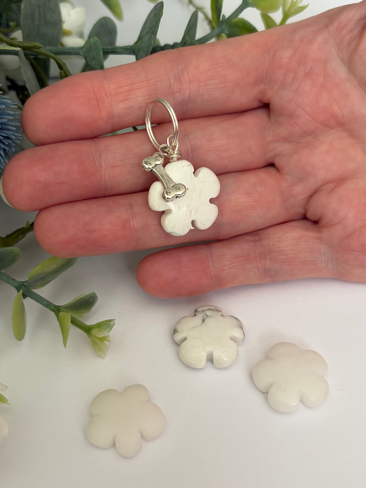 A hand holding a small white flower-shaped dog collar charm with a silver ring and bone detail, with three more matching flower charms scattered below on a white surface with green leaves in the background.