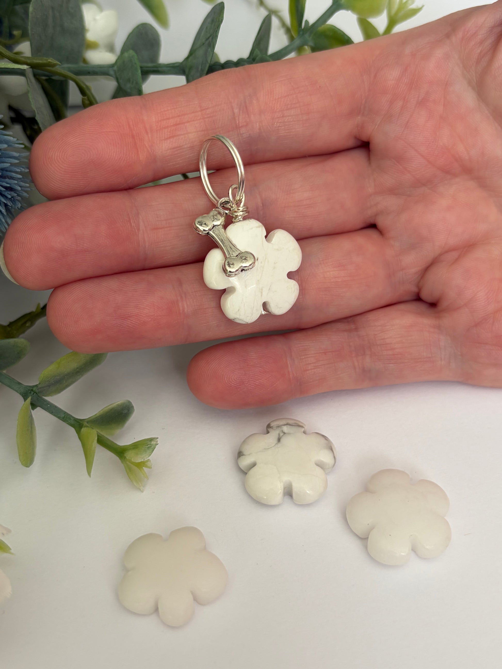 A hand holding a small white flower-shaped dog collar charm with a silver ring and bone detail, with three more matching flower charms scattered below on a white surface with green leaves in the background.