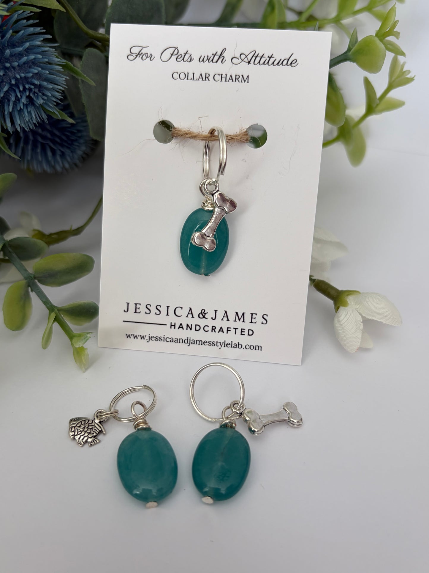 Teal and silver pet collar charm jewelry displayed on a white branded card, with matching drop earrings placed below and decorative greenery around the card.